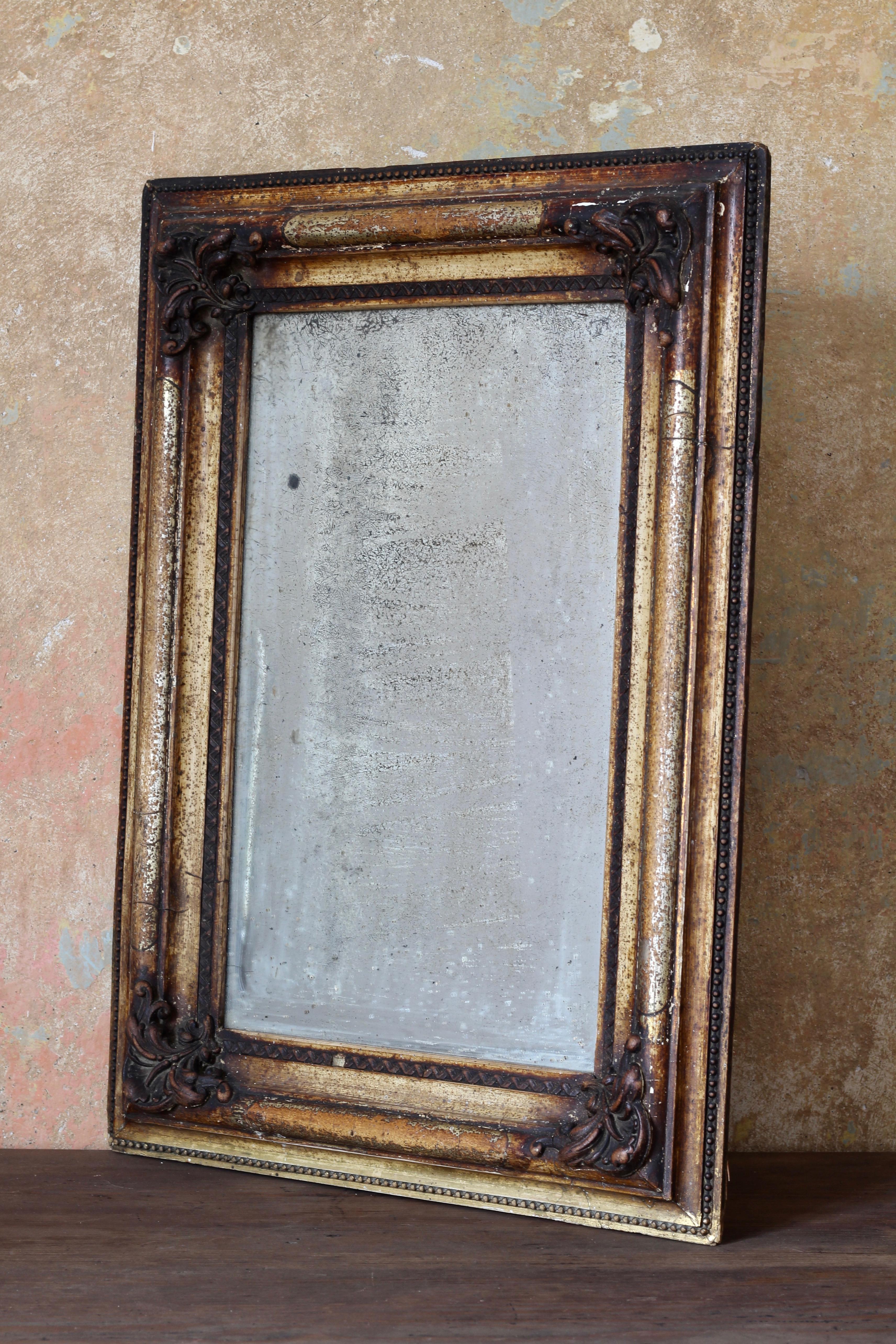 This wall mirror in original wooden frame has aged beautifully and acquired a unique patina over the time. The wooden frame in old-gold color has some beautiful decorative floral ornaments. The surface of the mirror must have seen it all-over the