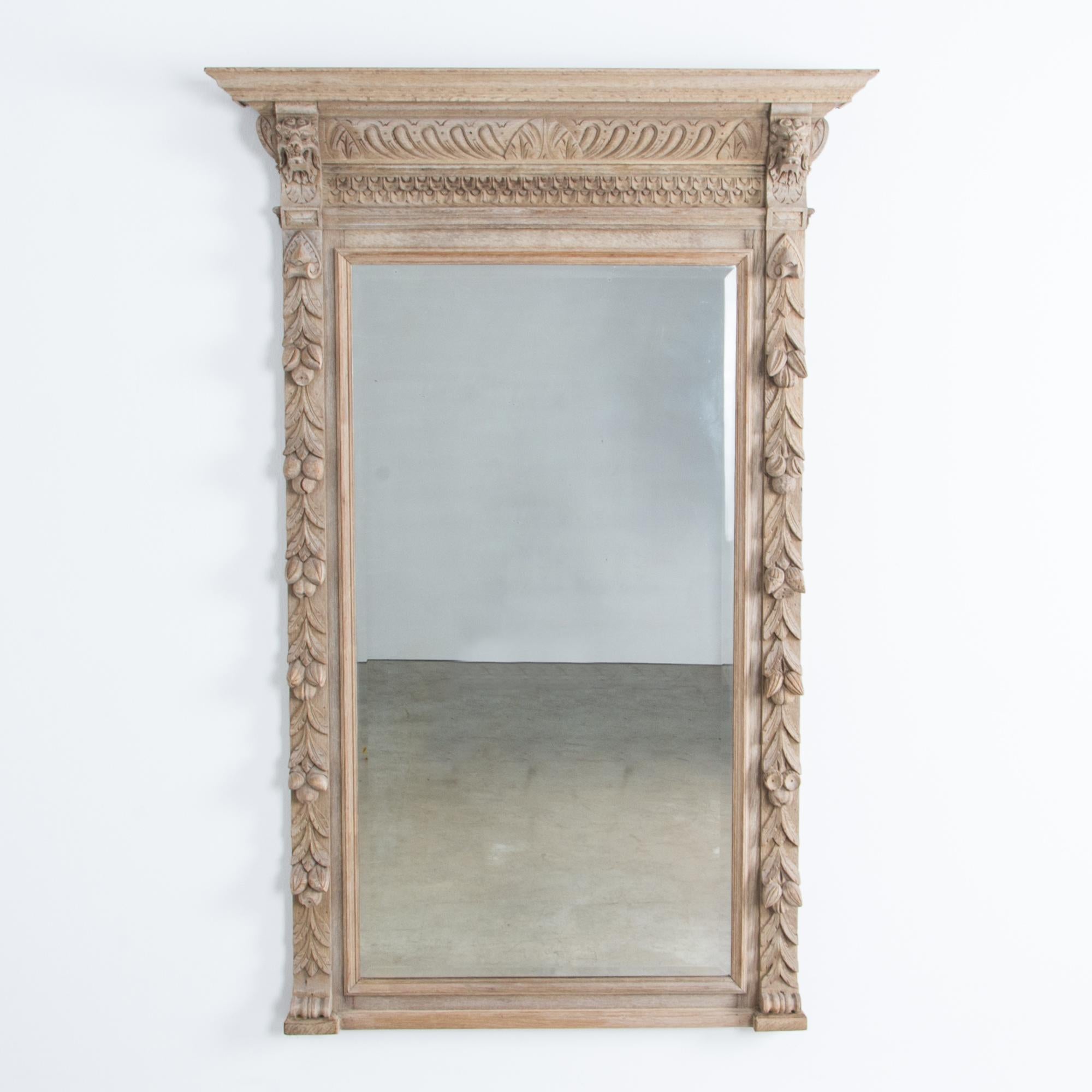 This antique wall mirror from Belgium, circa 1900, boasts an ornate carved wooden frame. The sides of the frame feature carved festoons of budding flowers, while from the corbels on either side emerge beautifully detailed carved lion heads. Both