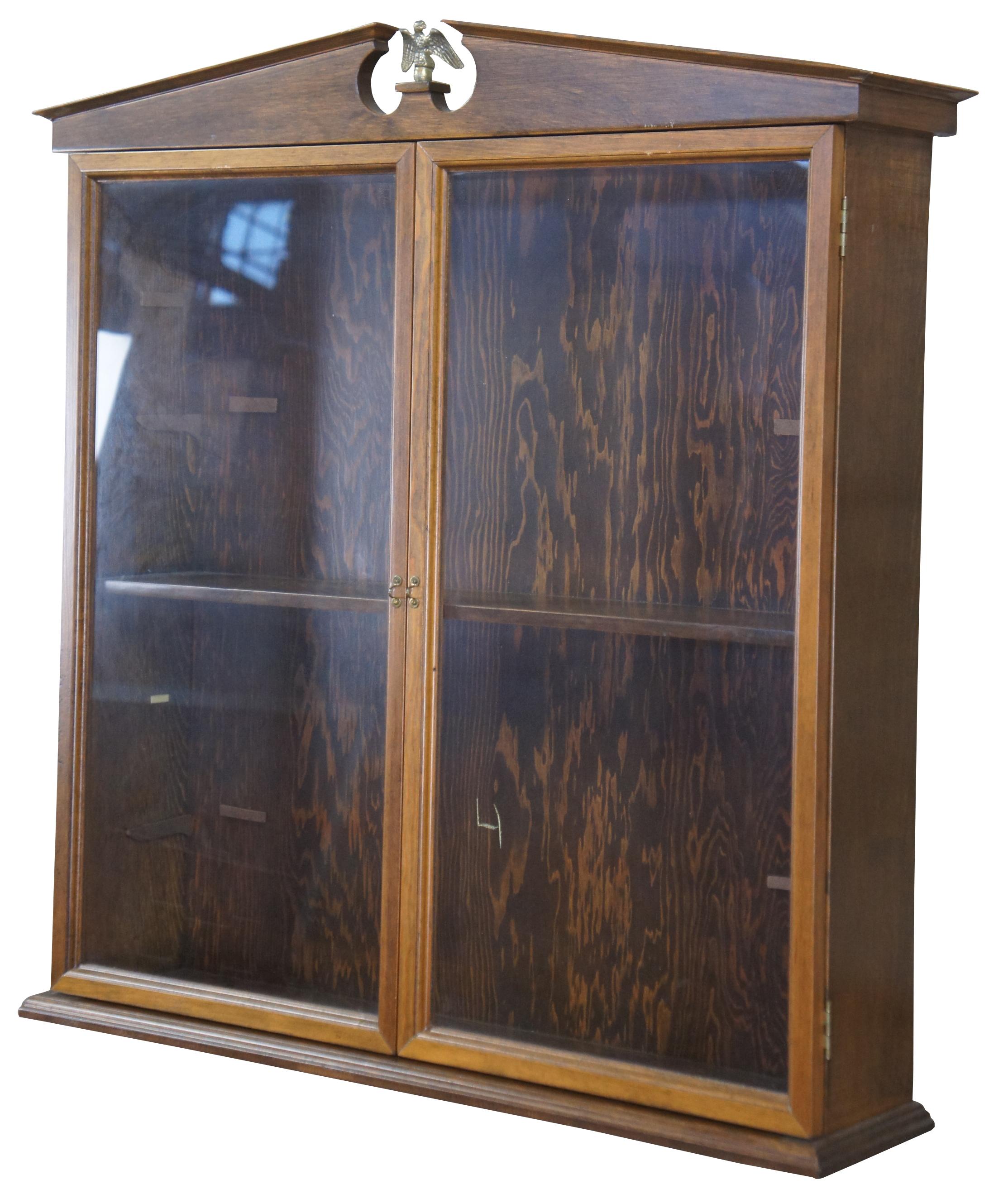 George III style wall curio or display cabinet, circa mid 20th century. Made of walnut featuring a broken pediment top, interior shelves with optional inserts, and glass doors locked by fish clasp. Two available. One has brass American Eagle finial,