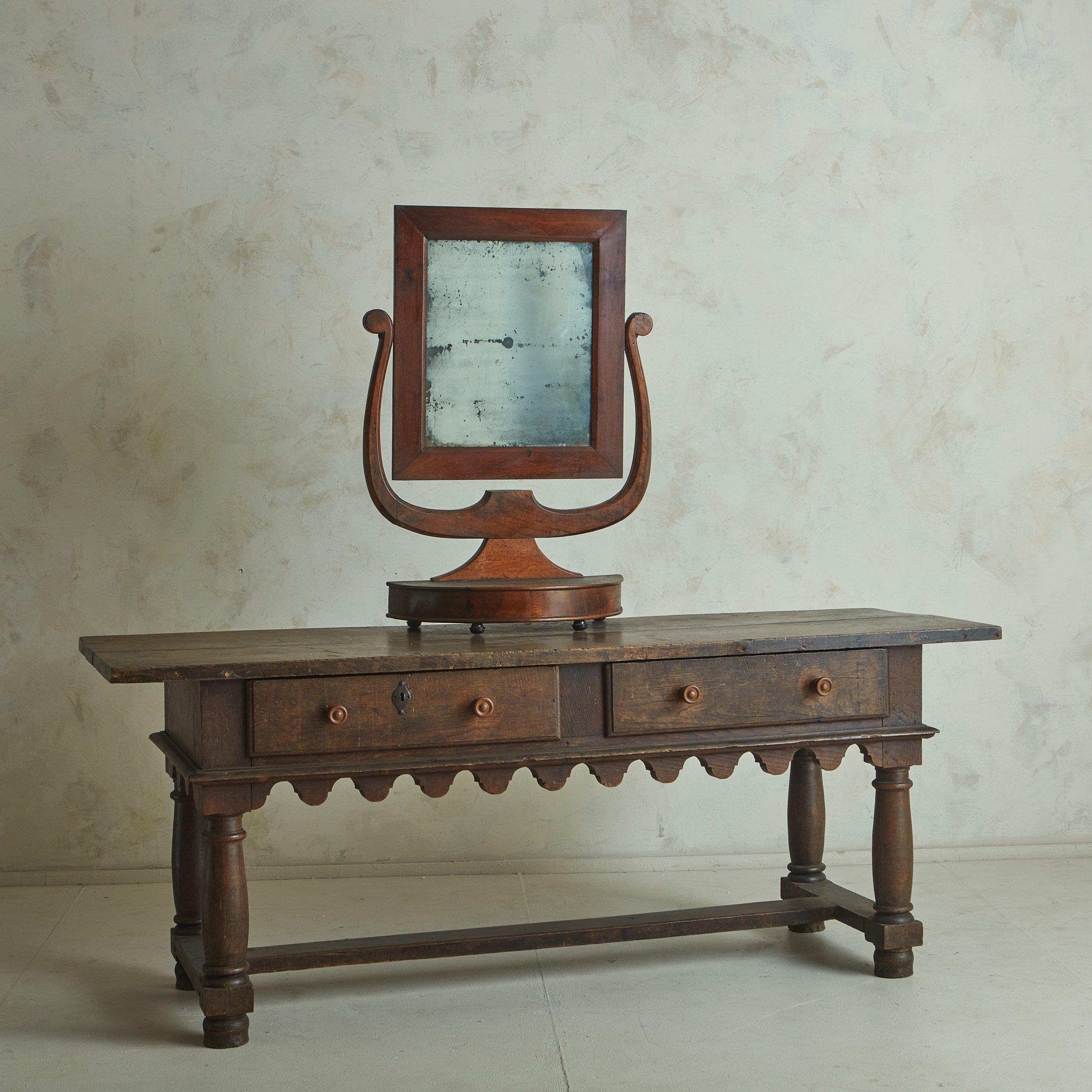 A handsome antique vanity mirror with a harp walnut frame and a demilune pedestal base. The frame supports a rectangular pivoting antiqued mirror and stands on four black metal feet. This piece has a gorgeous patina and features excellent