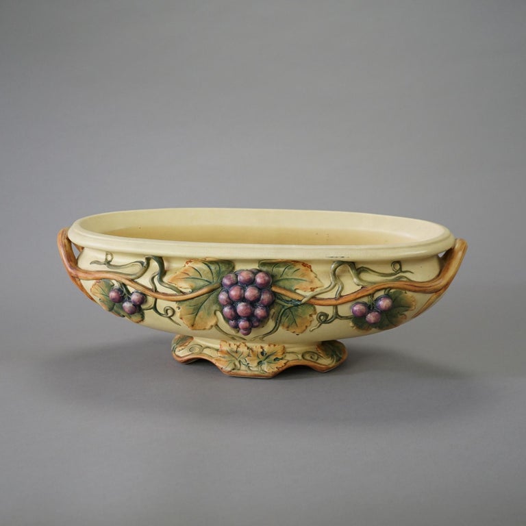 An antique bowl by Weller offers art pottery construction in oblong form with grape and vine in relief, double handles, and footed, en verso maker mark as photographed, c1930

Measures- 6.5'' H x 17.5'' W x 8.5'' D.
