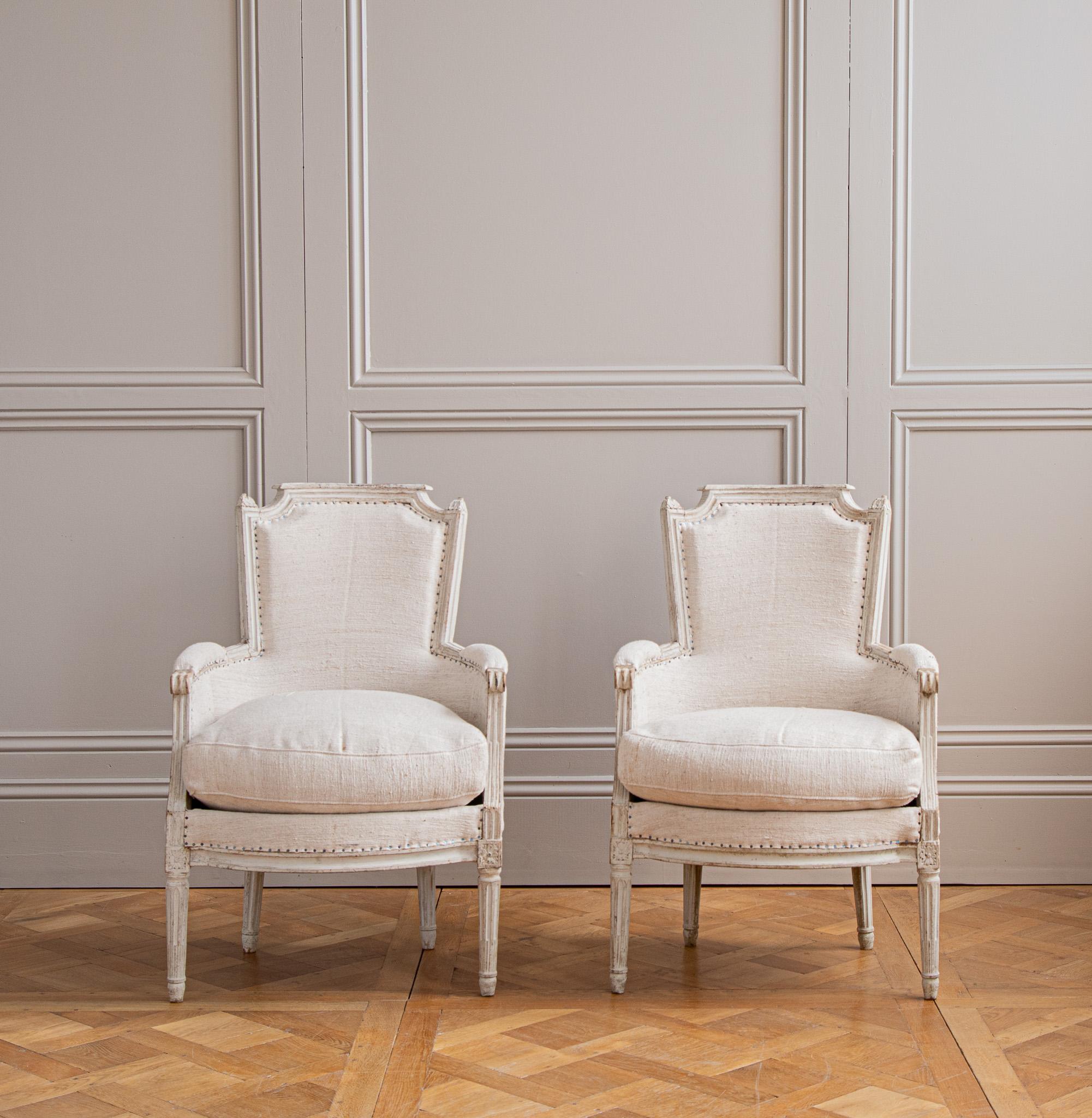 A pair of French Louis XVI style bergères in original paint from the 19th century. Rosettes on the corner posts with tapered and fluted legs. re upholstered in a Warm linen. 