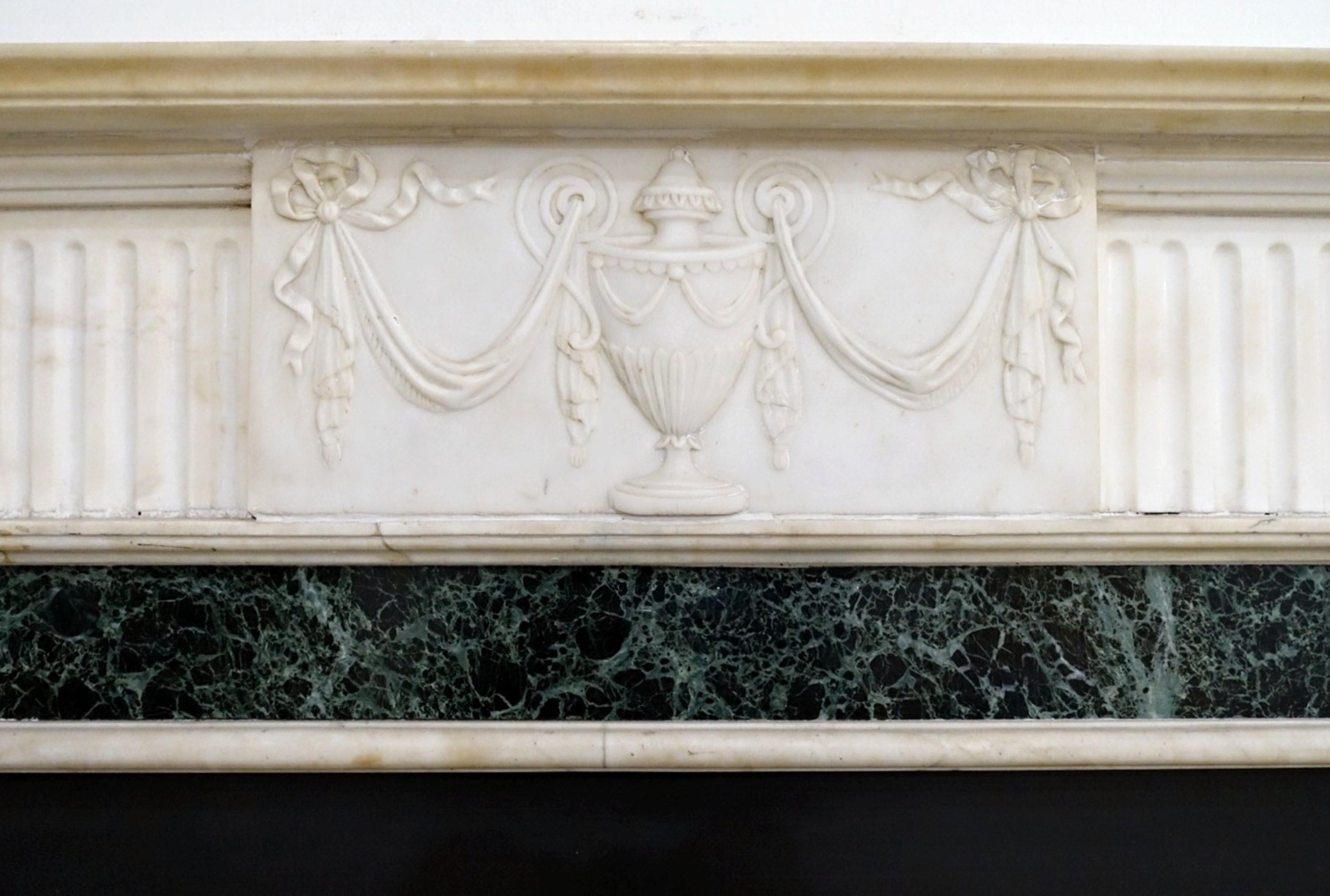 Presenting an elegant antique marble mantel graced by a skillfully carved urn centerpiece, adorned with oval motifs and fluted columns. The central structure is crafted from antique veined statuary marble, harmoniously complemented by a dark green