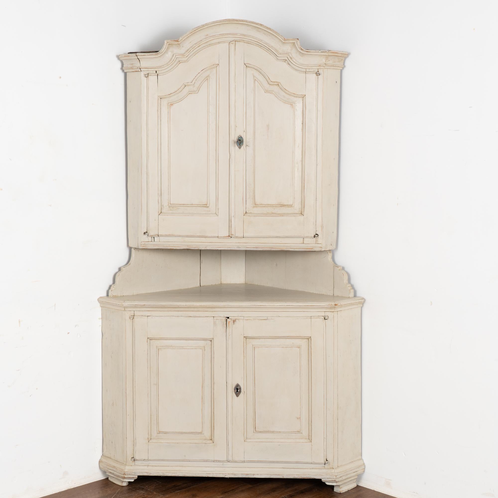 This pine corner cabinet has lovely paneled doors which compliment the curve of the bonnet or crown.
It is built in two sections and reaches 7' tall. 
The old white painted finish shows signs of distress, wear and some staining which simply add to