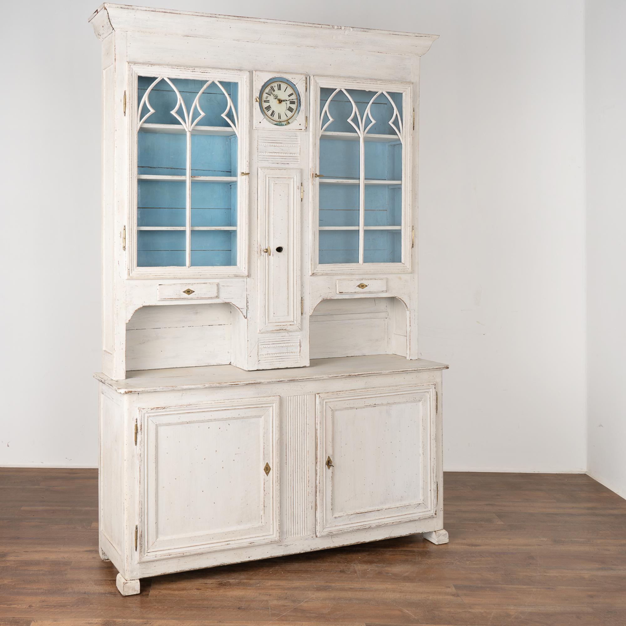 This delightful pine storage cabinet still displays the original clock face in the upper section with decorative carved molding accenting the glass doors.
The upper cupboard section has 3 removable shelves behind 2 glass doors and rests on the