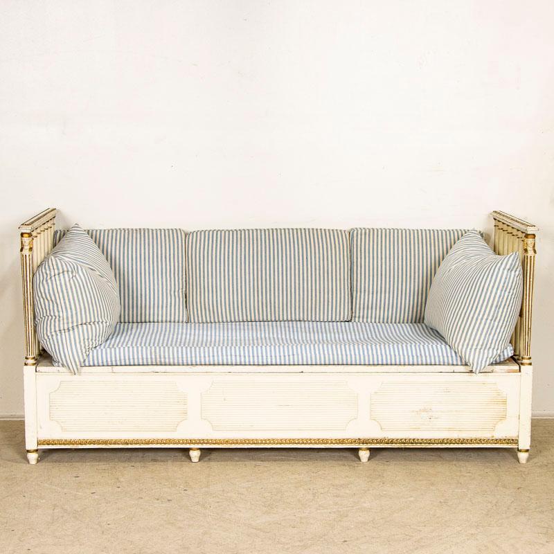 Painted white with gold accents, this antique Gustavian bench has a removeable cushion seat and open back. Architectural elements such as turned spindles create the sides and carved panels along the front add to the visual intrigue of this