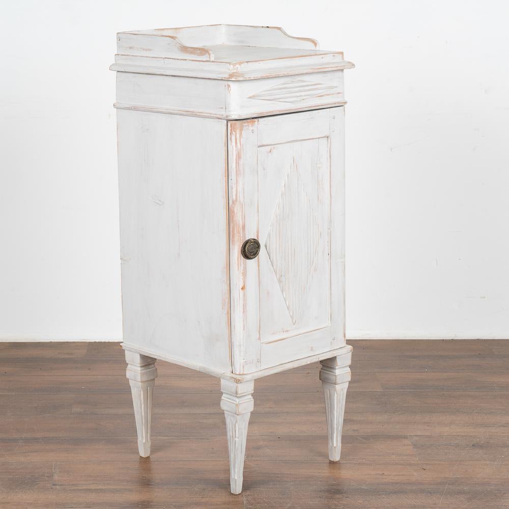 Small Swedish country white painted nightstand or side table standing on fluted tapered feet.
Traditional diamond motif on single panel door which opens to reveal an interior drawer and shelf.
Newer professionally applied white painted finish