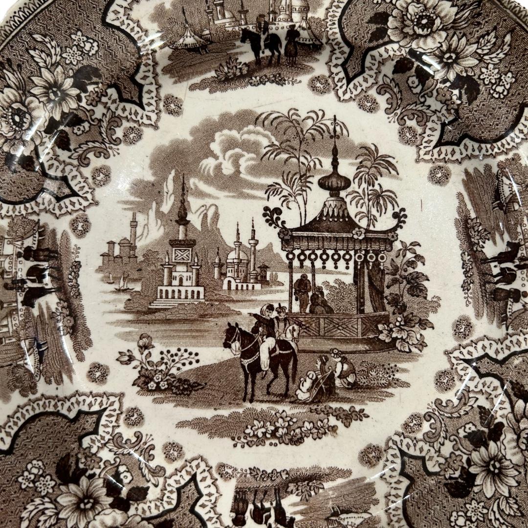 This elegant ceramic plate from William Adams & Sons made in Staffordshire, England was made during the mid 19th century. It has a scalloped edge with flowers and scenes of gazebo and temples in center. The plate, “Palestine” pattern, is in