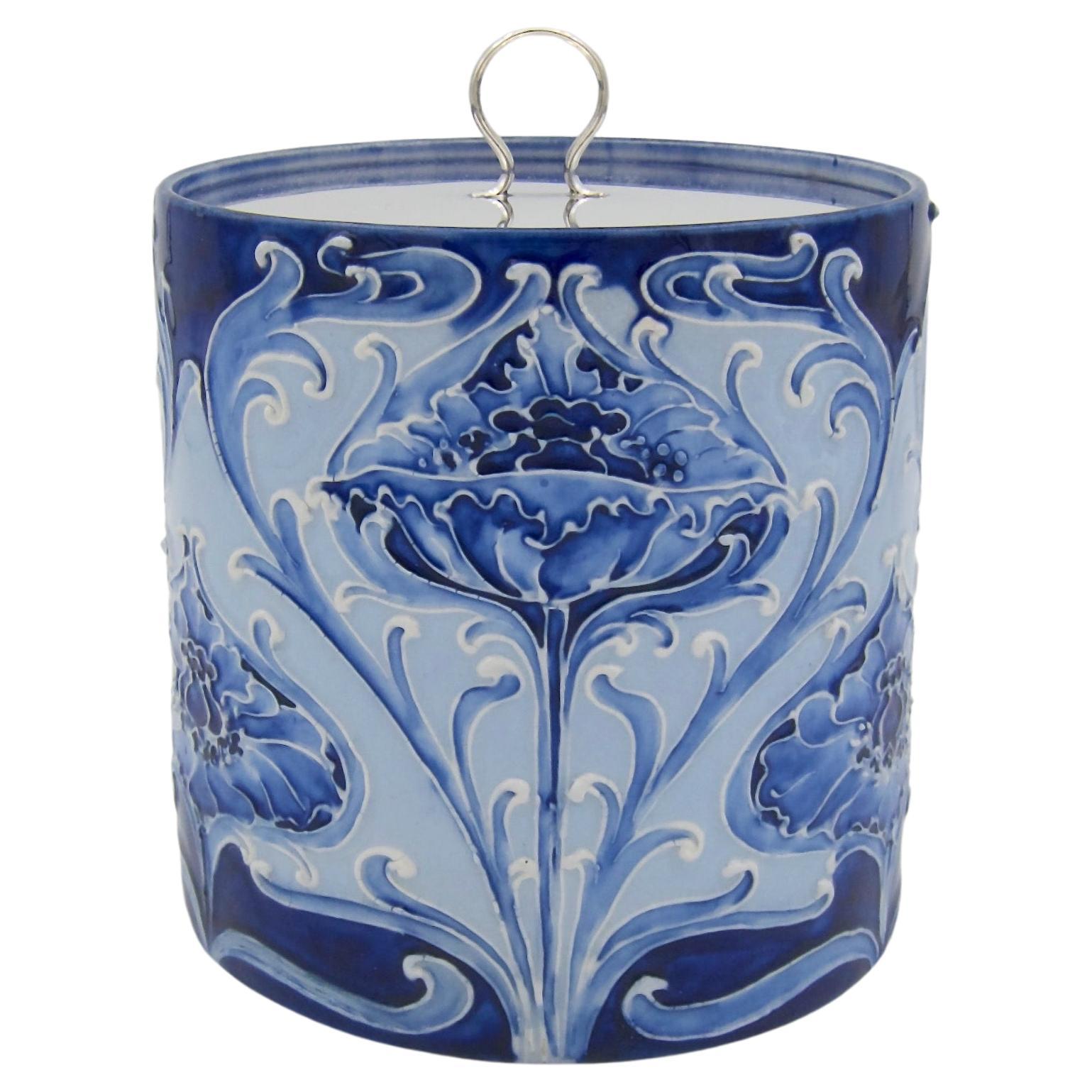 A signed William Moorcroft Florian ware art pottery biscuit barrel with stylized floral decoration in shades of blue and a silver-plated lid. The canister was made in England for James Macintyre & Co., Ltd. of Washington Works, Stoke-on-Trent,