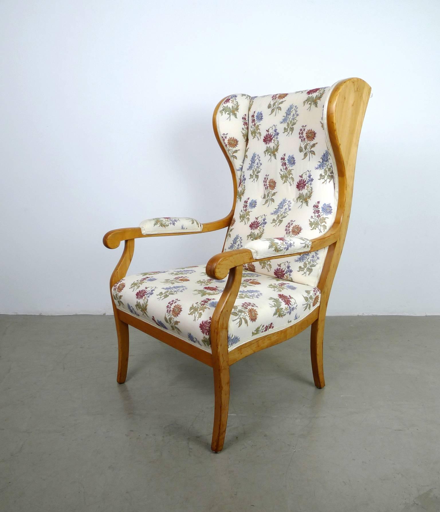This wingback chair features a solid cherry frame and a bright fabric upholstery cover with floral motifs. The backrest is also covered on the back with fabric. The armrests are partially upholstered on the right and left. The fabric surfaces are