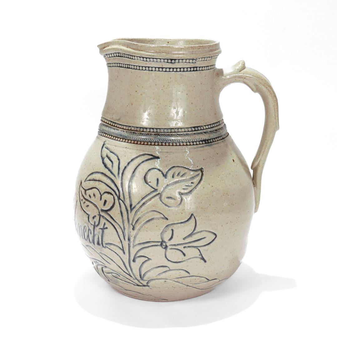 A fine antique Bristol glazed stoneware pitcher.

By the Wingender Pottery of Haddonfield, NJ.

Of ovoid form with a narrow footed base, an incised & dated inscription and floral (tulip) decoration to the body, a molded foliate handle, and a coggled