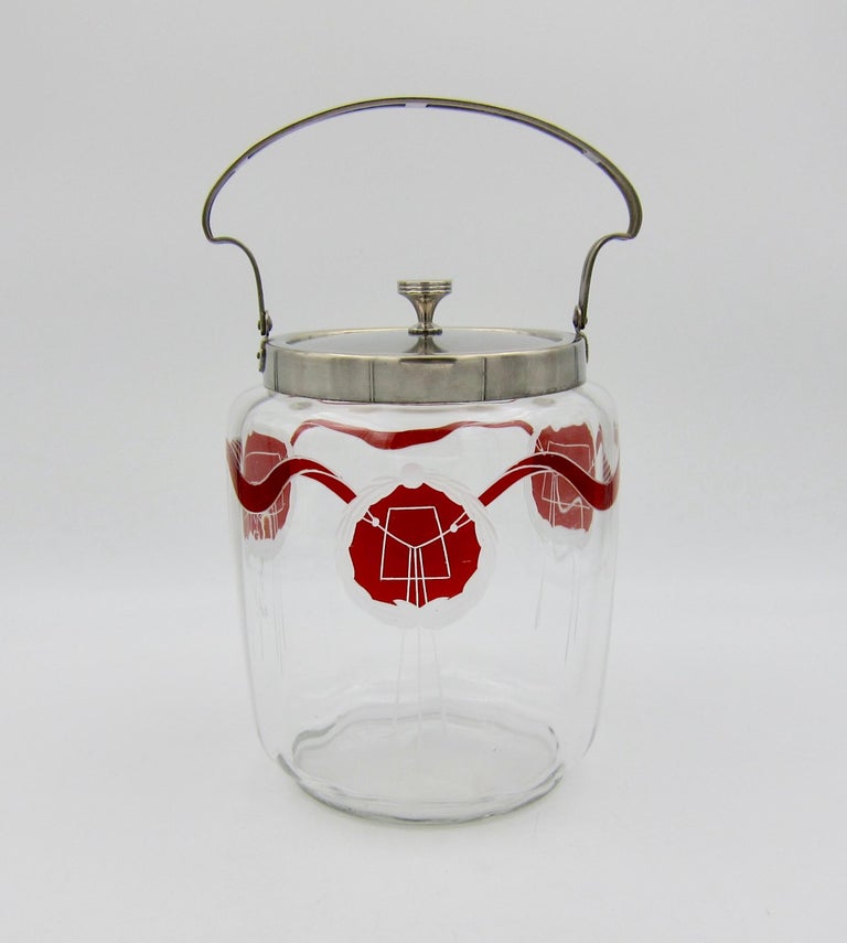 An antique biscuit barrel jar from Württembergische Metallwarenfabrik (WMF) of Germany. The early 20th century covered jar has a silver plated metal rim, bail handle, and lid. The clear glass body is decorated with Secessionist / Art Nouveau décor