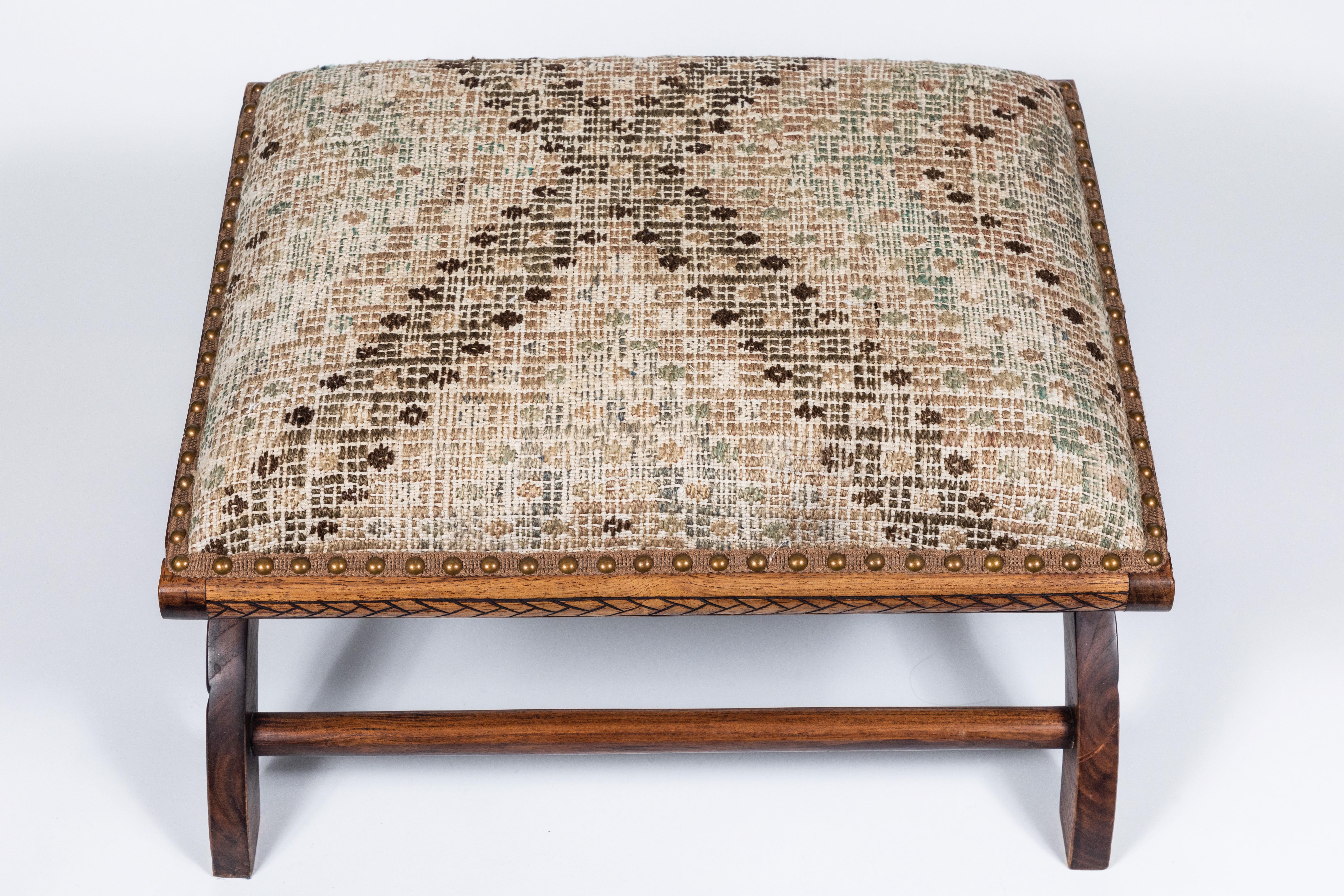 Antique wood footstool with carved flowers on the side, upholstered in a vintage Turkish rug with nailhead trim.