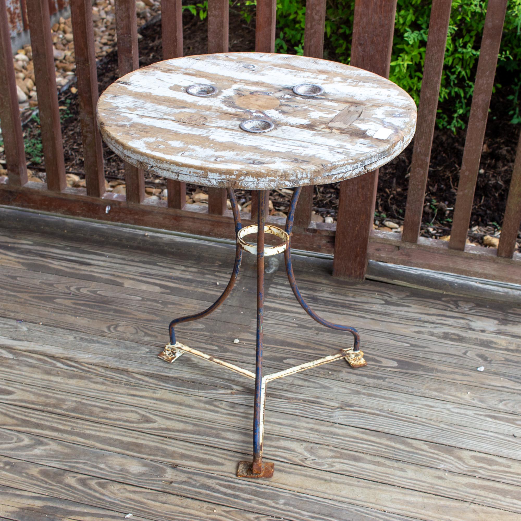 This charmingly rustic wood & metal bistro table was sourced in Spain. The top is wood with three round metal rivets which are attached to the metal base. The wood has a distressed painted finish with the remnants of white paint, also echoed on the