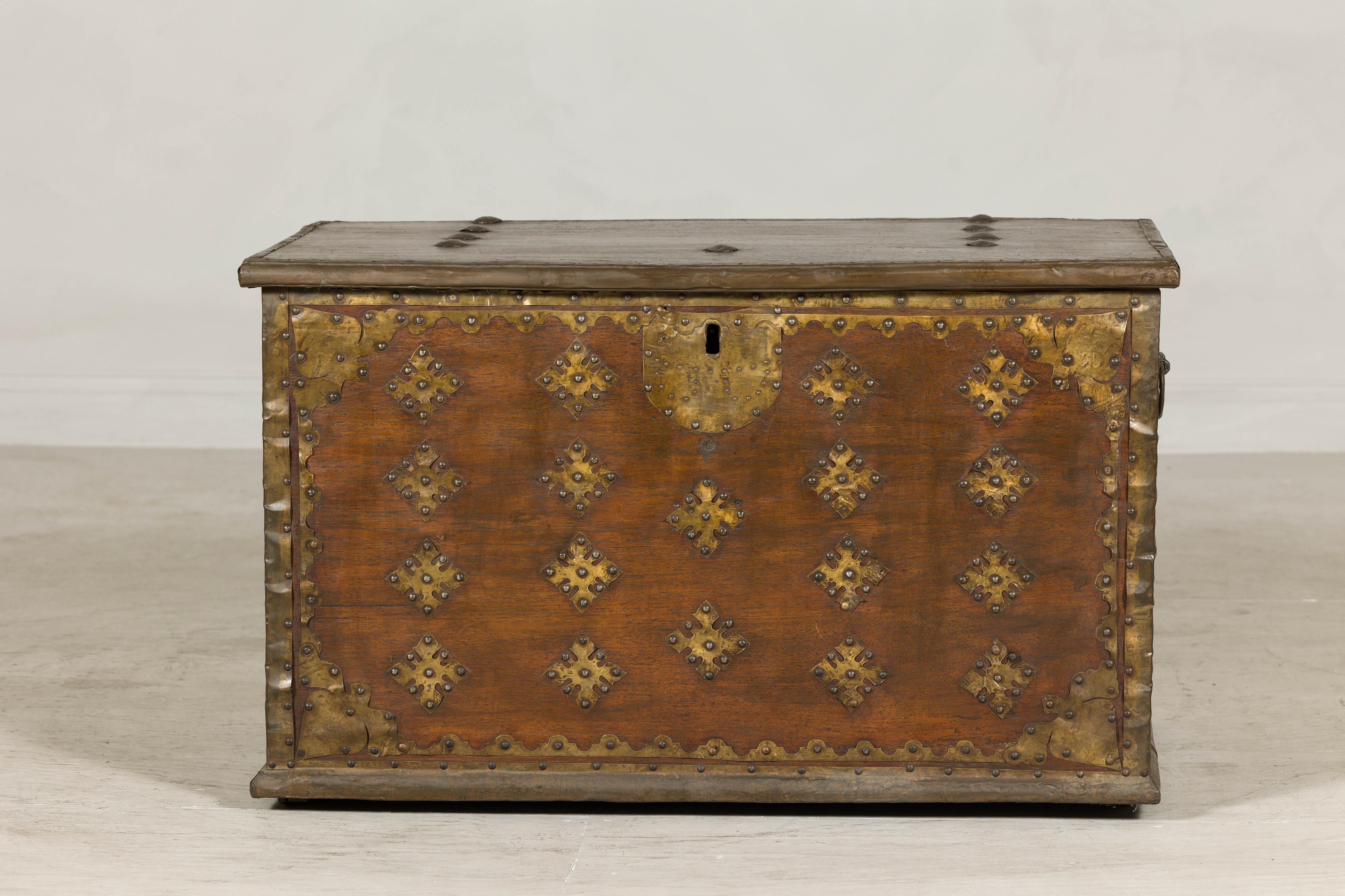 This large antique trunk is distinguished by its bold metal appliqué, presenting a striking blend of practical craftsmanship and ornamental appeal. Constructed of hardwood and reinforced with hand-hammered brass straps and studs, the piece is built