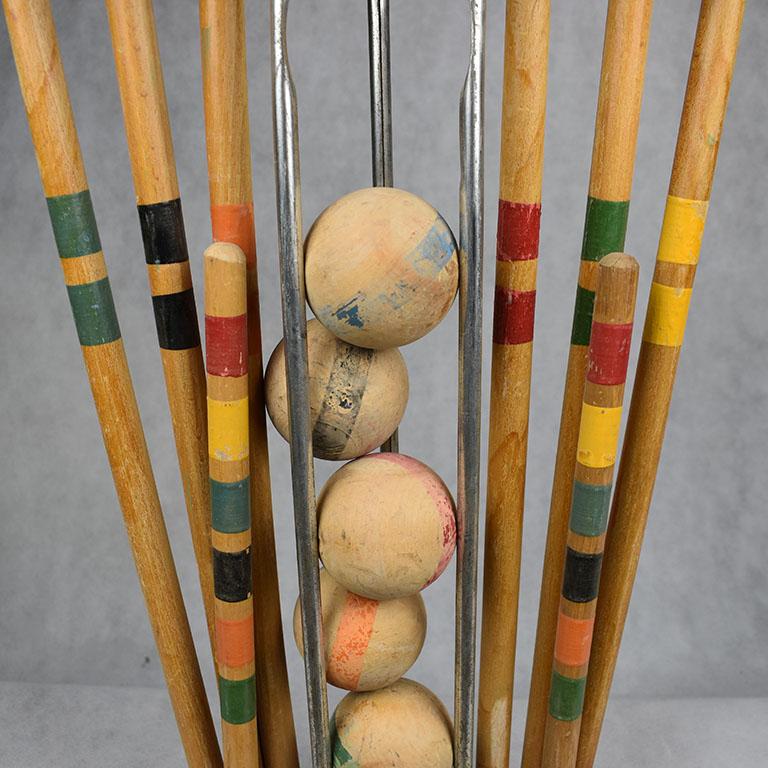 Antique set of croquet mallets, balls and stakes. Painted in yellow, green, red, orange, black and blue. The set sits in a green stand with metal handle. The balls sit comfortably between the clubs and the wooden stakes are held, just like the