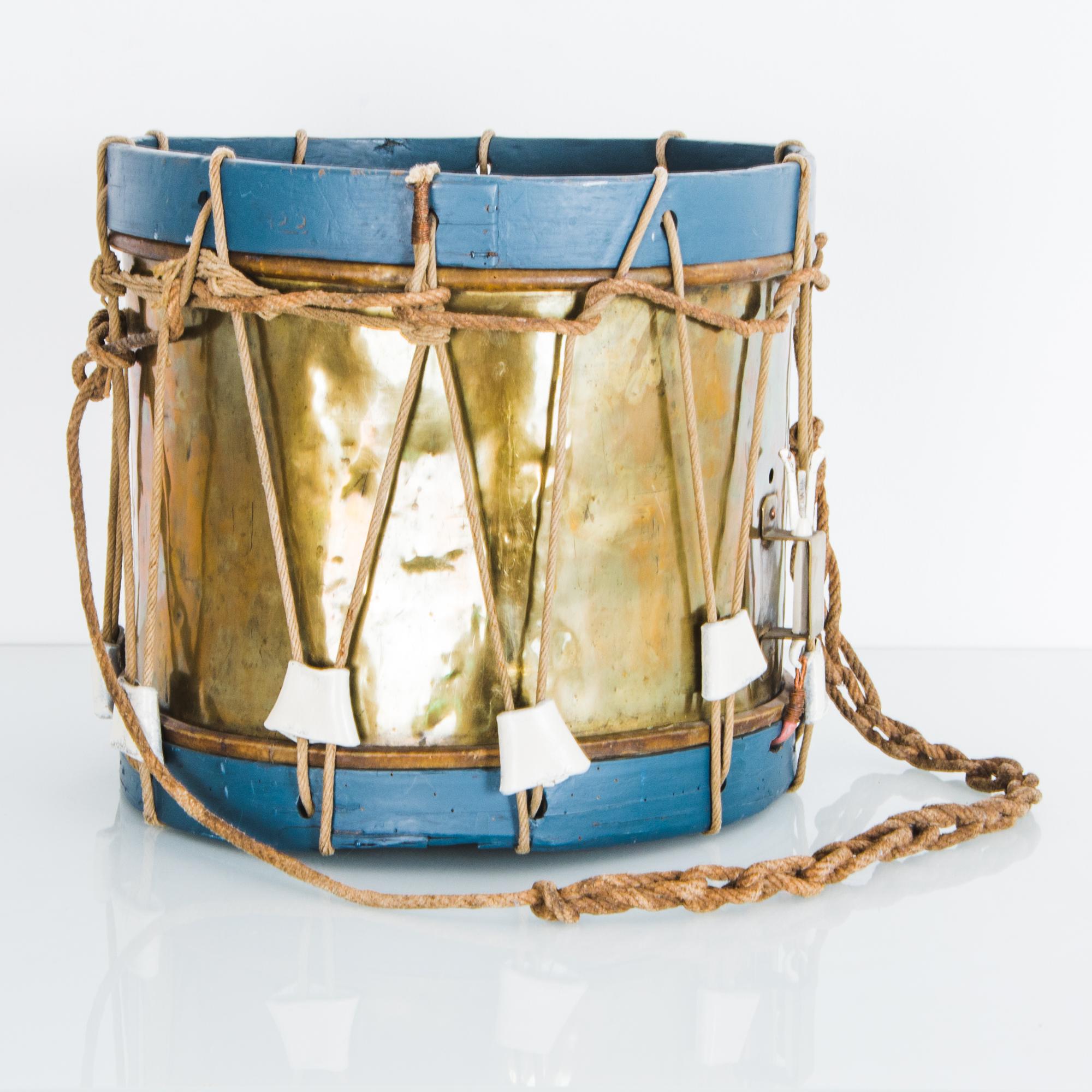 This antique drum hails from the United Kingdom, circa 1920. The shell of the drum is made of golden metal, while the wooden rims are painted a cheerful contrasting blue. The light weathering of drum-skin and drum-strap gives an antique charm to