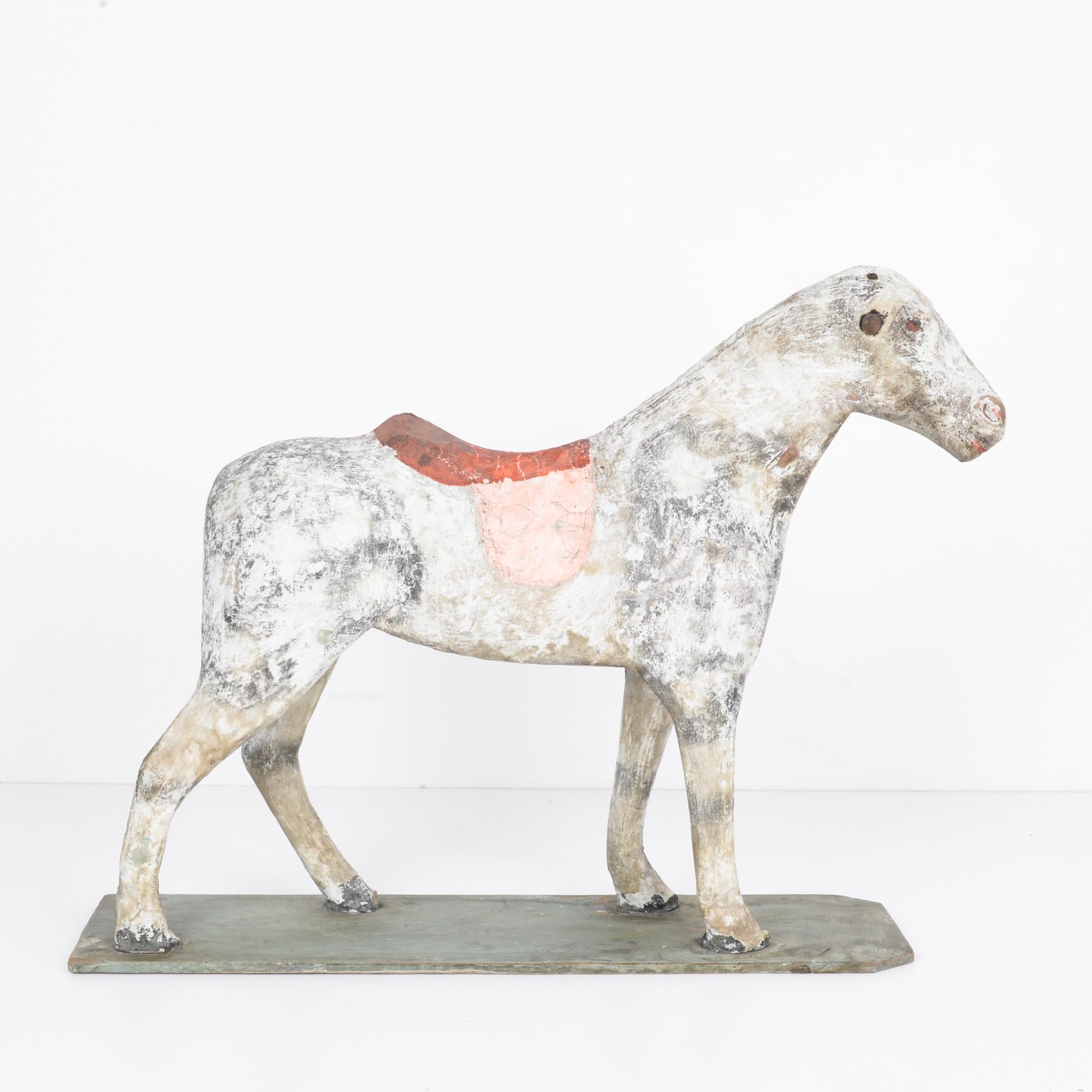 A wooden horse from France, circa 1900. Originally painted white, the finish has weathered into a striking patina, scuffed and textured, with shades of pigeon gray and sepia. Red-painted accents — the saddle, mouth and nostrils — create a bright