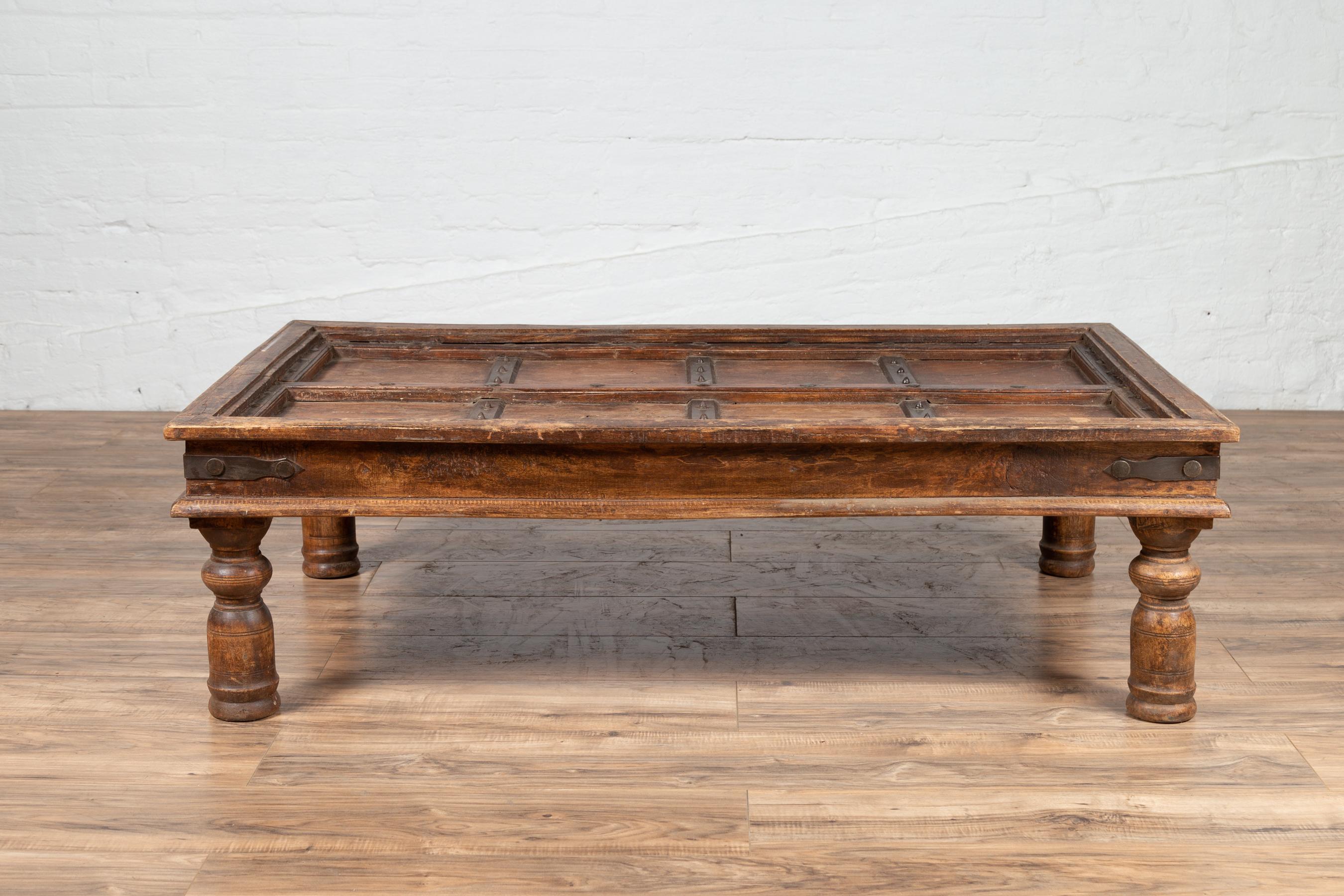 An antique Indian palace wooden door made into a coffee table with iron accents and baluster legs. Born in India during the early years of the 20th century, this exquisite palace door has been made into a coffee table. The top is rhythmically