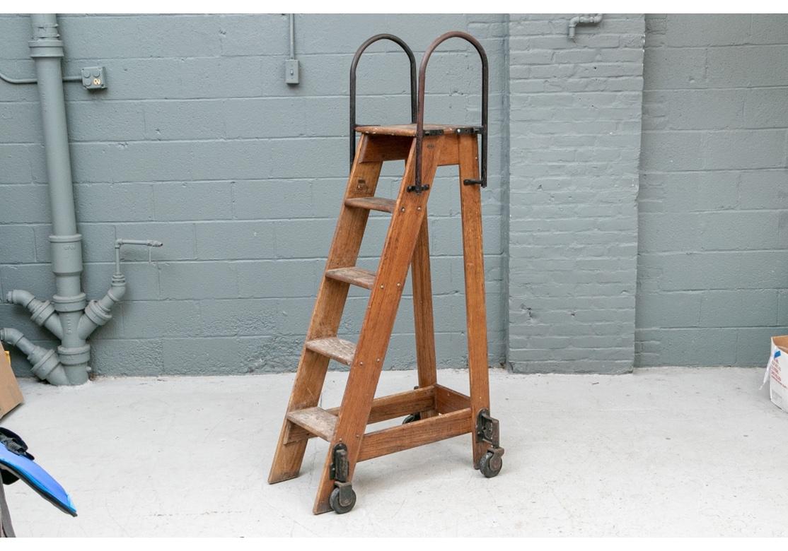 A five rung Industrial Era Ladder with a pair of tall arch shaped iron handles on top. The base with black painted iron mounts and wheels. Original Label and Oval Number Plate is affixed. 
Measures: height 67