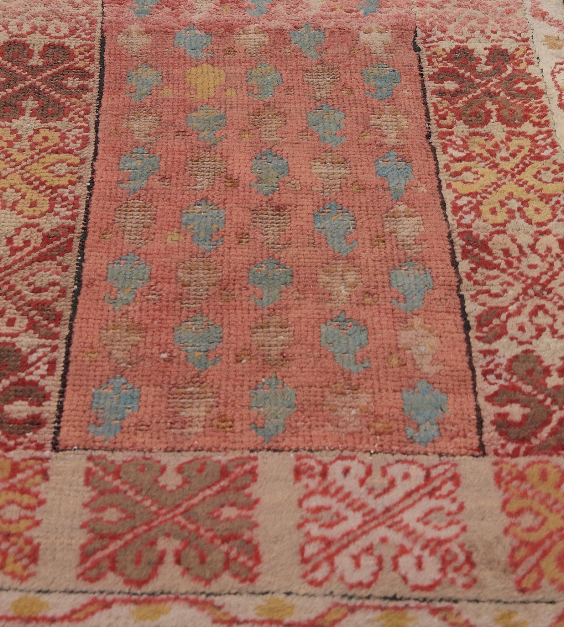 This antique Oushak runner has a coral-red field with diagonal rows of light blue and ivory boteh, in a broad ivory border of shaded soft brown and golden yellow alternating hooked floral panels, outer ivory ribbon-pattern vine stripe, plain outer