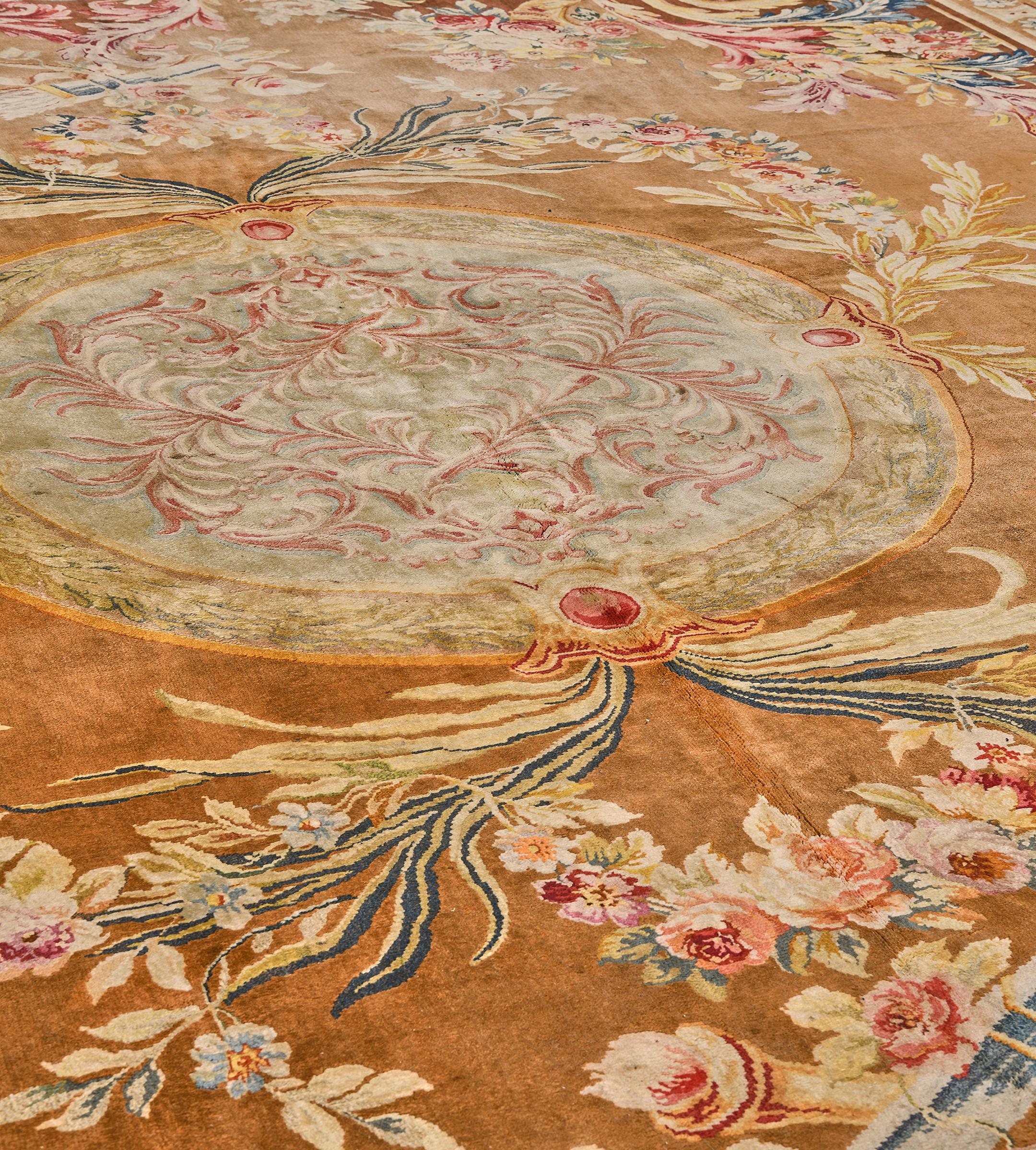 This antique, circa 1900, French Savonnerie rug has a golden-brown field with a central eau-de-nil roundel containing a shaded ivory and red interlocking acanthus leaf vine, an outer soft green laurel-leaf band with a pairs of leafy sprays issuing
