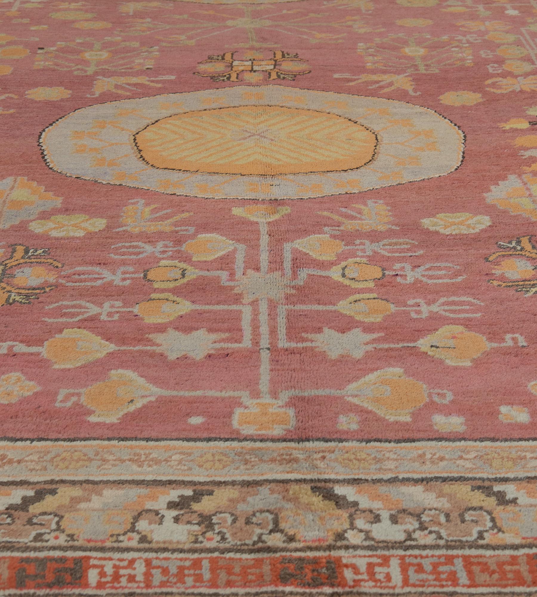 This antique Khotan rug has a shaded raspberry-red field with scattered angular floral stems and flowerheads around a pair of roundels with a central apricot lozenge and a golden-yellow angular serrated vine surrounded by a broad light-blue band
