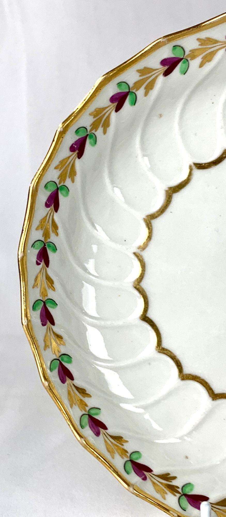 Questo elegante piatto in porcellana di Worcester della fine del XVIII secolo è scanalato e decorato lungo il bordo con una bella serie di foglie dipinte in verde chiaro, viola e dorato.
La semplice decorazione rivela la scanalatura della