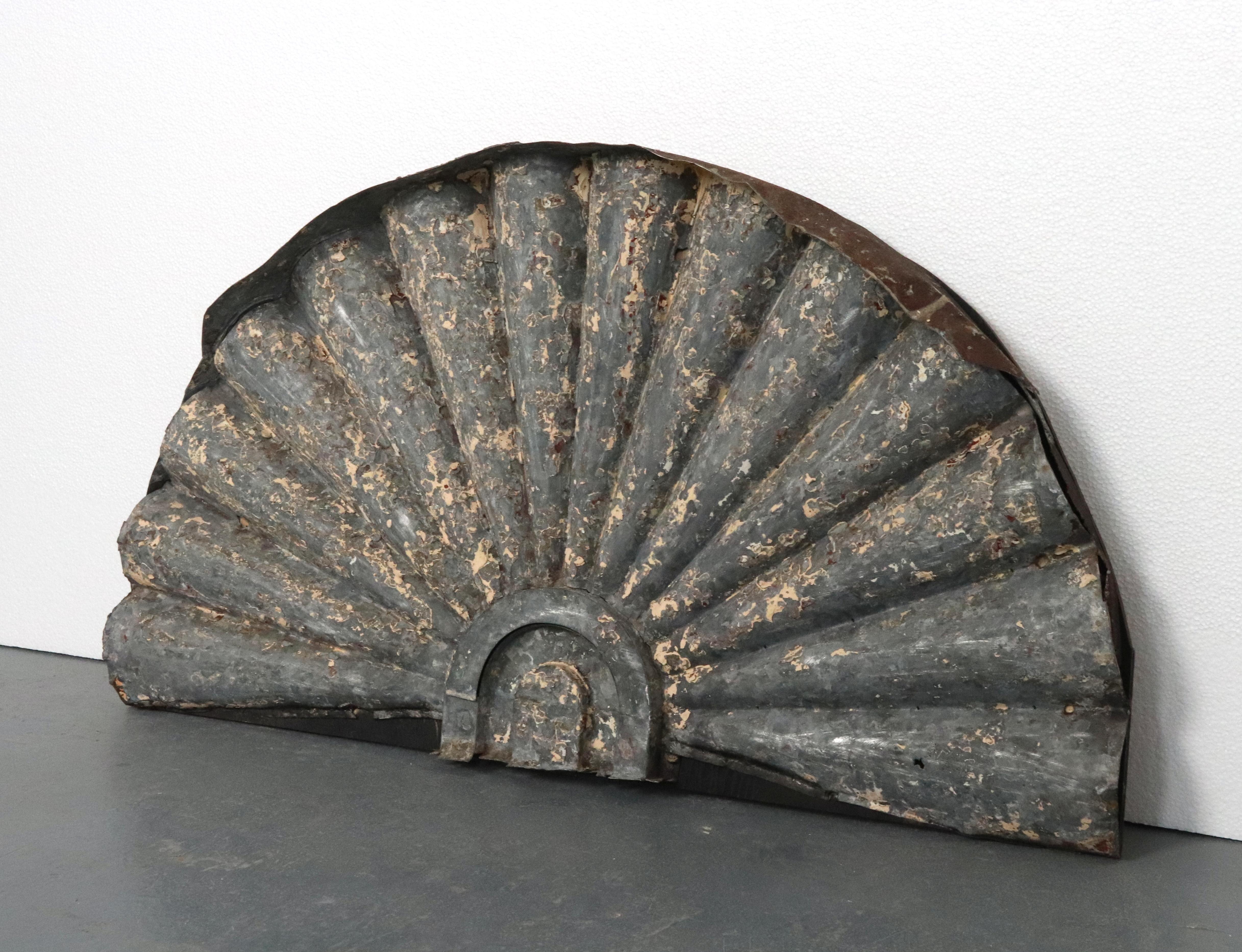 Elemento arquitectónico de zinc recuperado que presenta una serie de segmentos estriados que crean una textura ondulada, parecida a la forma de un abanico estilizado o un arco. Este elemento se fijó a una pieza de madera para exponerlo como obra de
