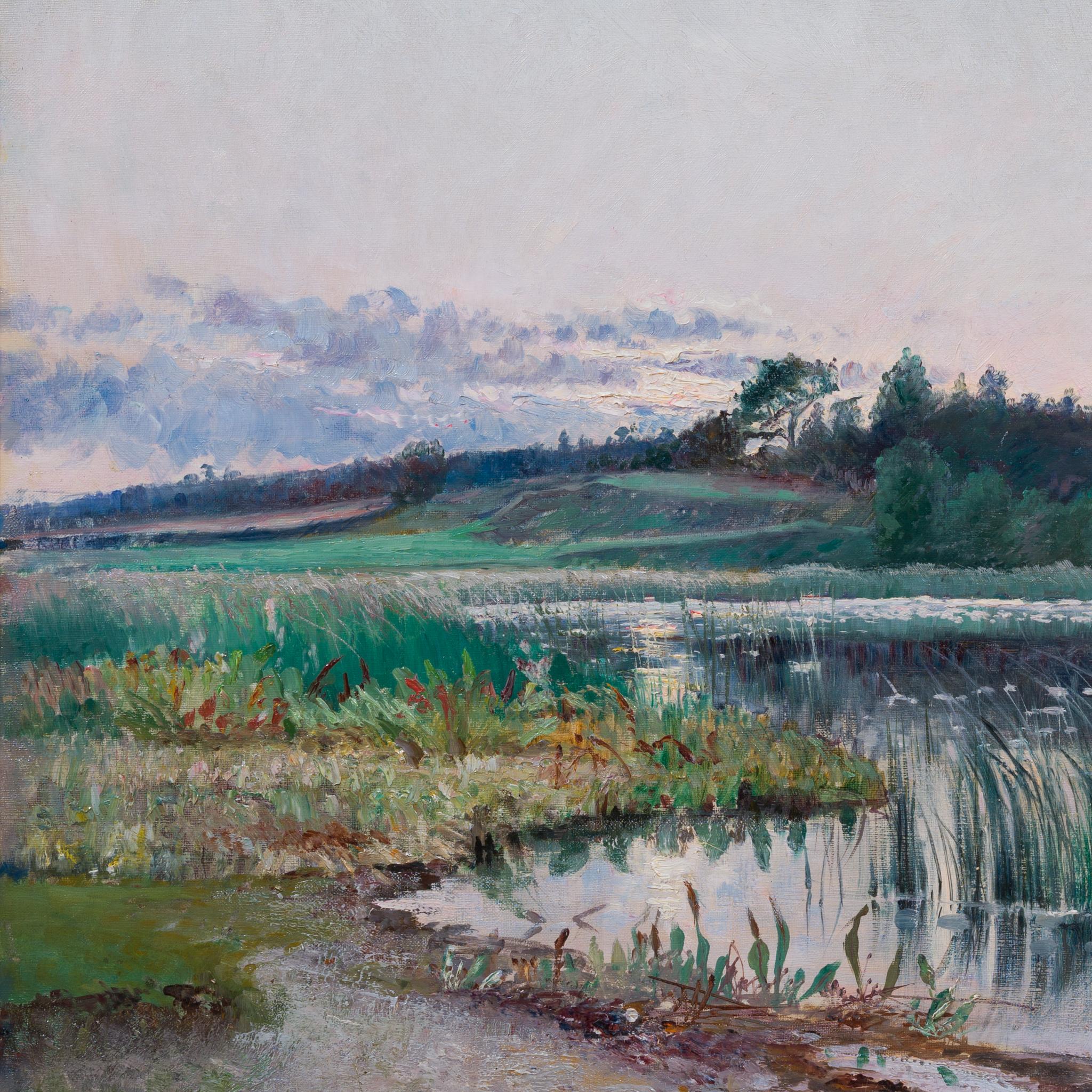 In the soft evening light, a lone figure rows a small boat across the calm water, while tall reeds fringe the quiet shoreline in the foreground. Rolling wooded hills embrace the lake in the distance, their forms gently silhouetted against a sky