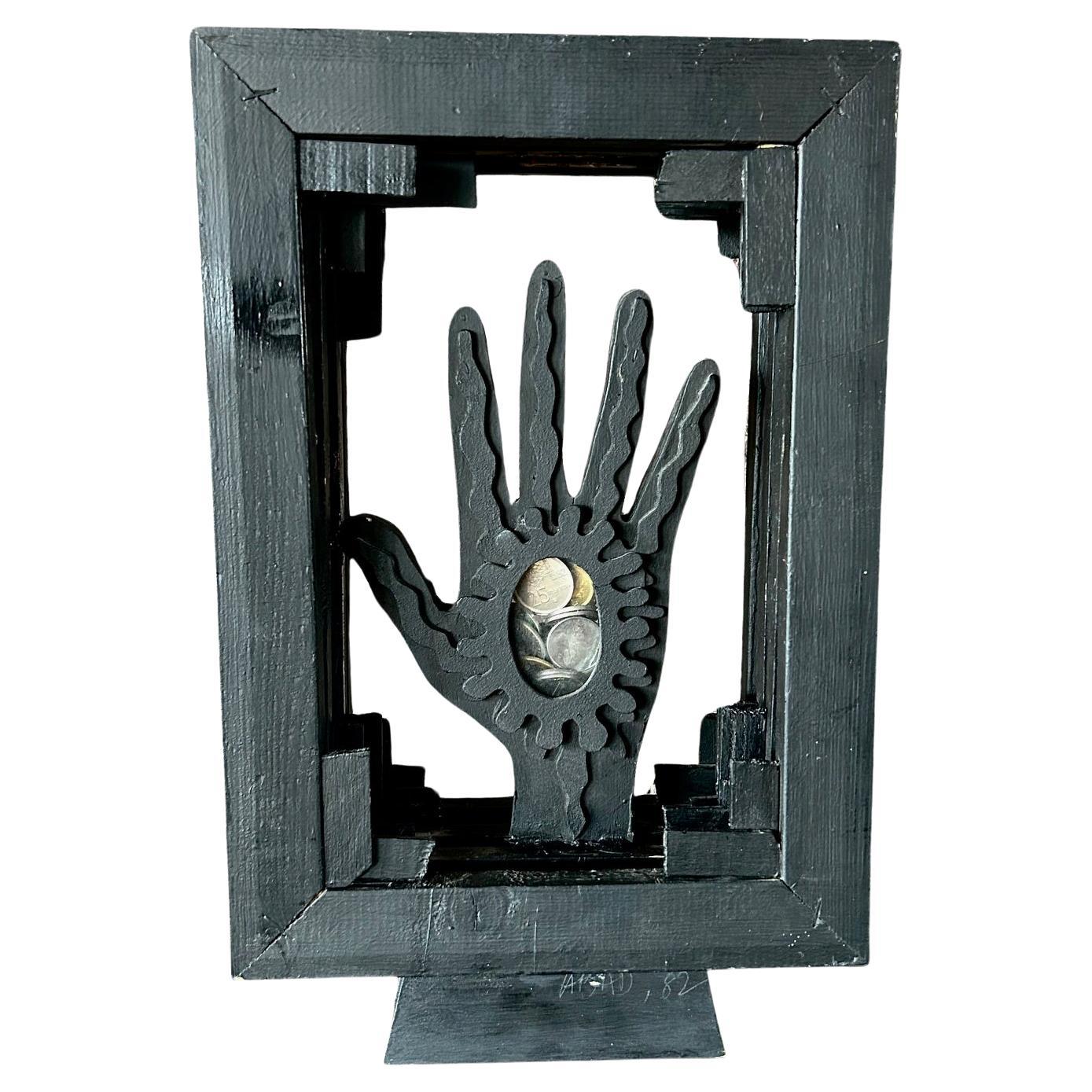 Wooden Sculpture in black featuring a central hand within a framework and containing metal coins by Spanish artist Antoni Abad. 
Abad's works have been exhibited in major museums and galleries worldwide.