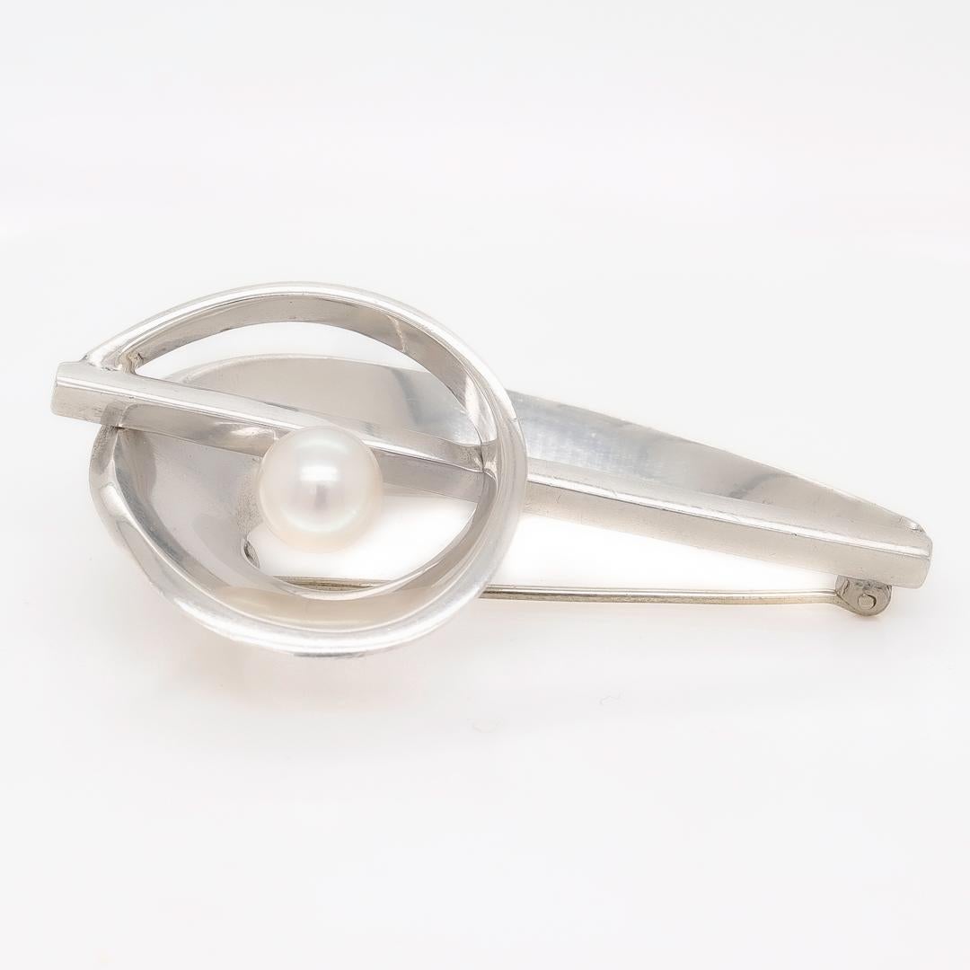 A fine Modernist silver brooch.

In sterling 970 silver. 

By Antonio Pineda.

Comprised of looping silver bands reminiscent of a bass clef with a striking accent pearl.

Marked to the reverse for Taxco Mexico / 970 / Antonio Pineda's crown mark /