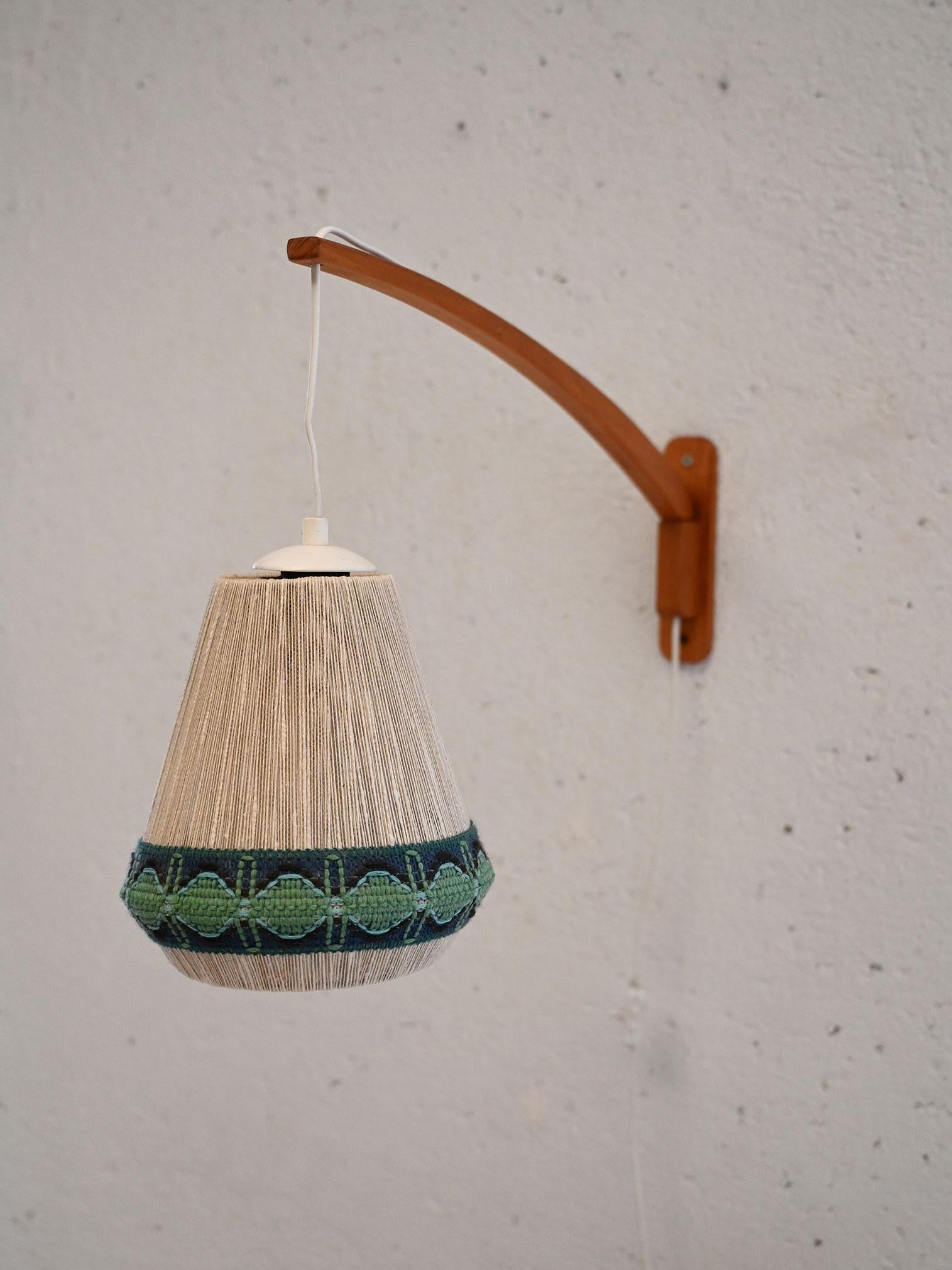 1960s teak wall sconce with decorated fabric abat-jour and adjustable wire. Elegant and functional design.

Scandinavian design wall lamp from the 1960s, featuring a teak base and an ornate fabric abat-jour with green and black geometric patterns.