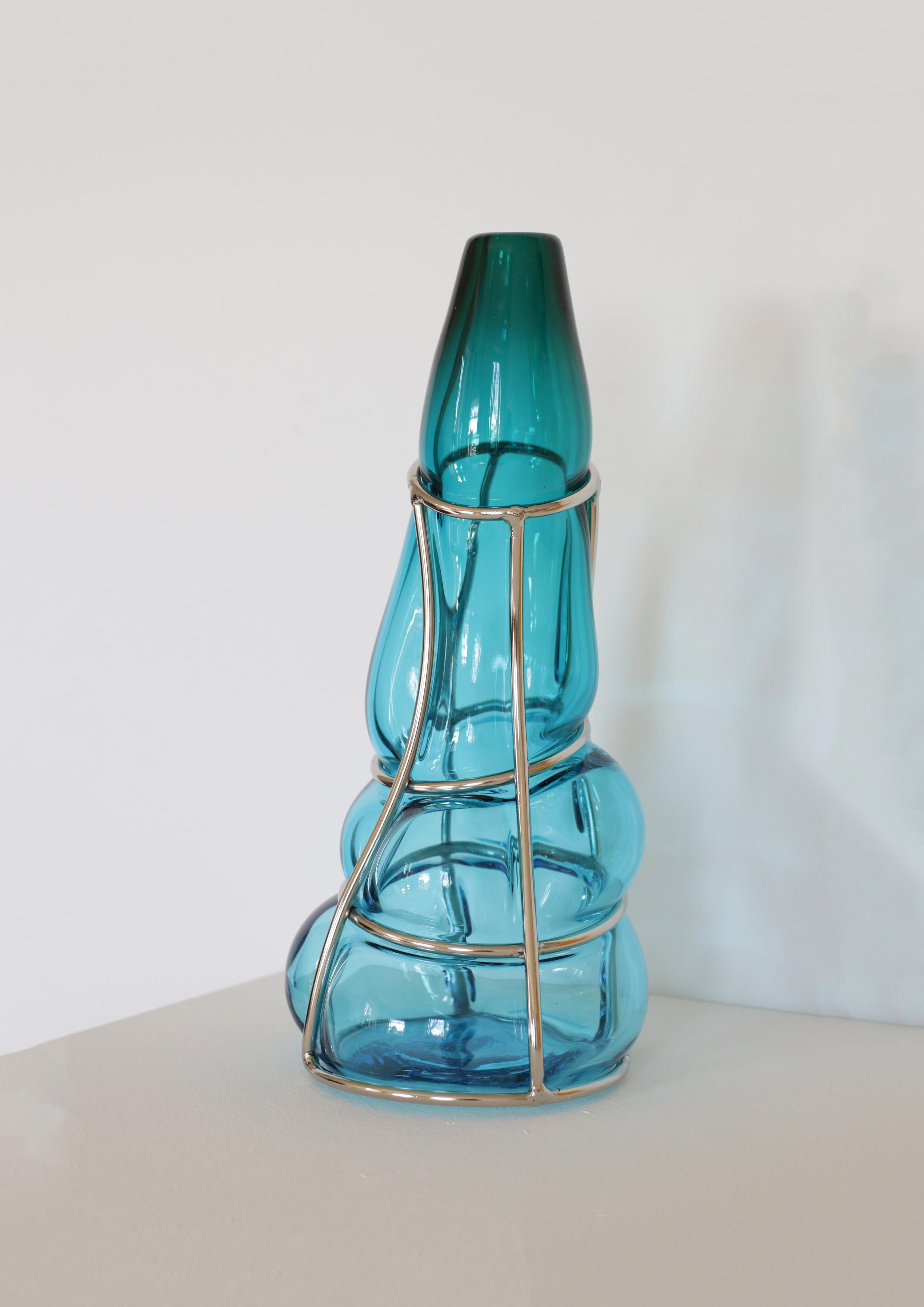 Vaso scultura Aqcuamare Babà di Alice Crepaldi
Un pezzo unico.
Dimensioni: P 16,5 x L 16,5 x H 36 cm.
Materiali: Vetro di Murano e metallo.

Adornato da uno stelo leggermente più corto ma elegante, questo vaso ingabbiato utilizza la tecnica dello