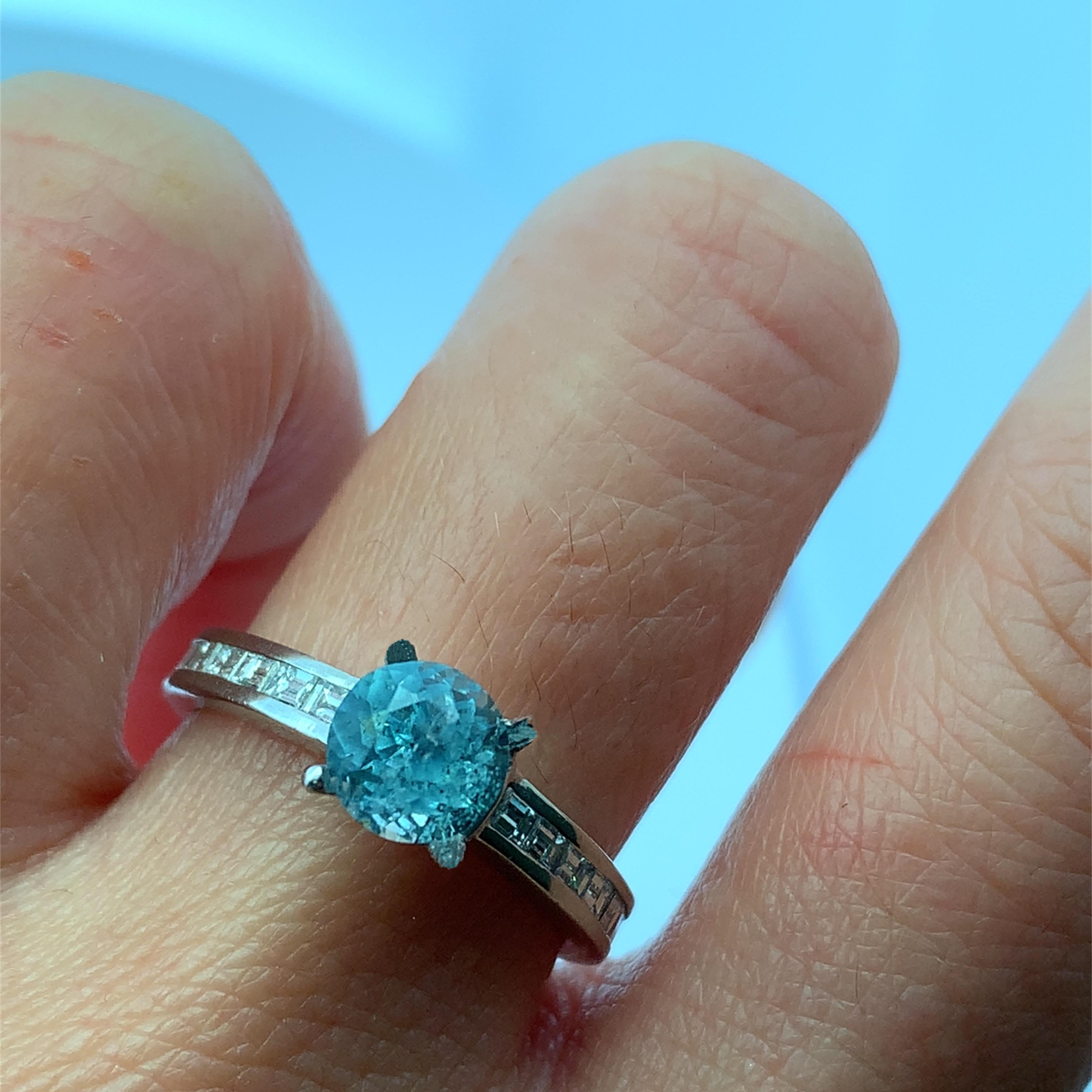 Scopri la squisita bellezza di questo elegante anello con gemme di acquamarina blu e diamanti. Caratterizzato da uno splendido centro  L'acquamarina da 1,25 carati a taglio rotondo circondata da diamanti baguette da 0,69 carati G VS1, è un pezzo
