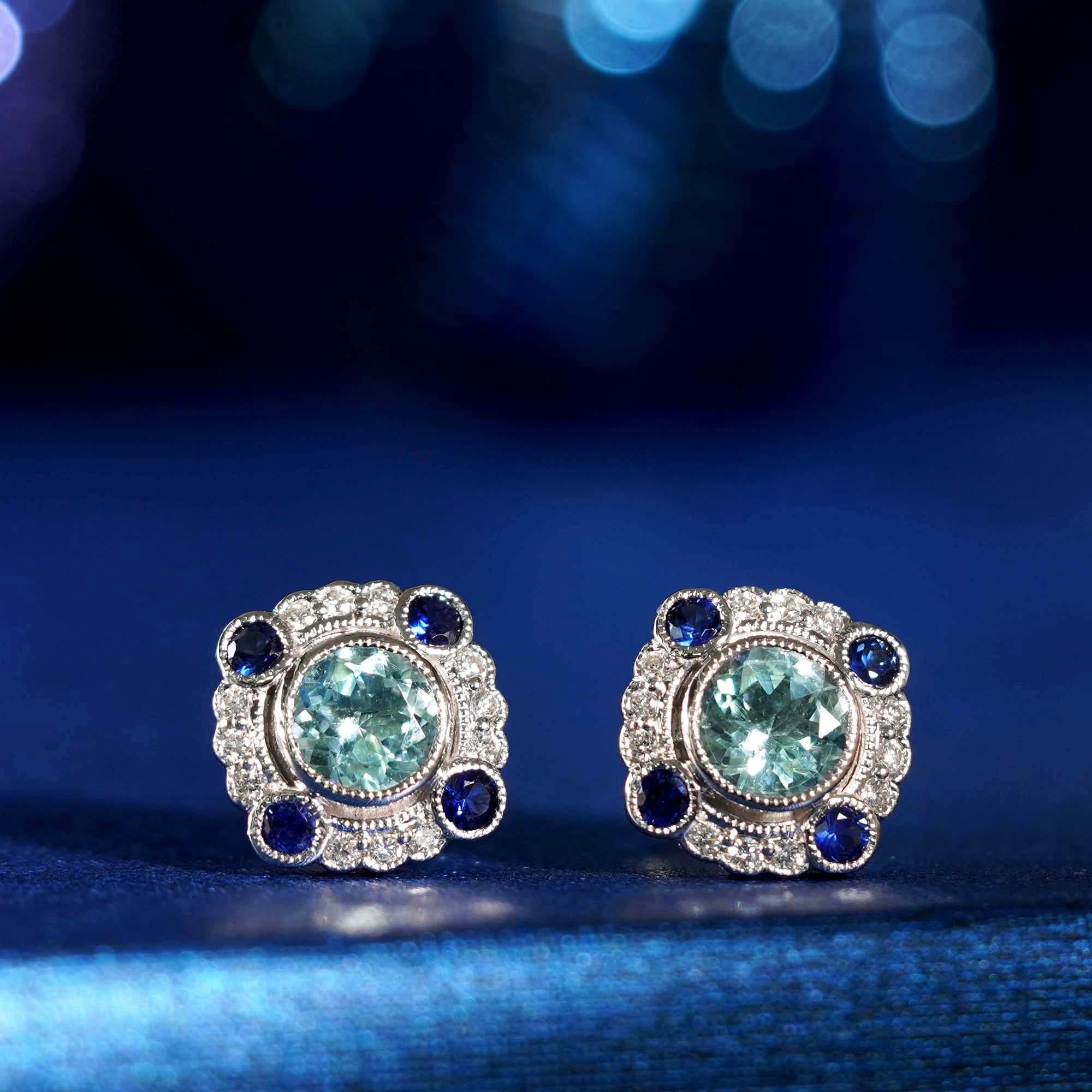 A radiant round aquamarine at the center, encircled by a sparkling diamonds. Four round blue sapphires accent the top, bottom, left, and right, adding bold contrast and geometric elegance. Inspired by classic Art Deco design, these earrings offer a