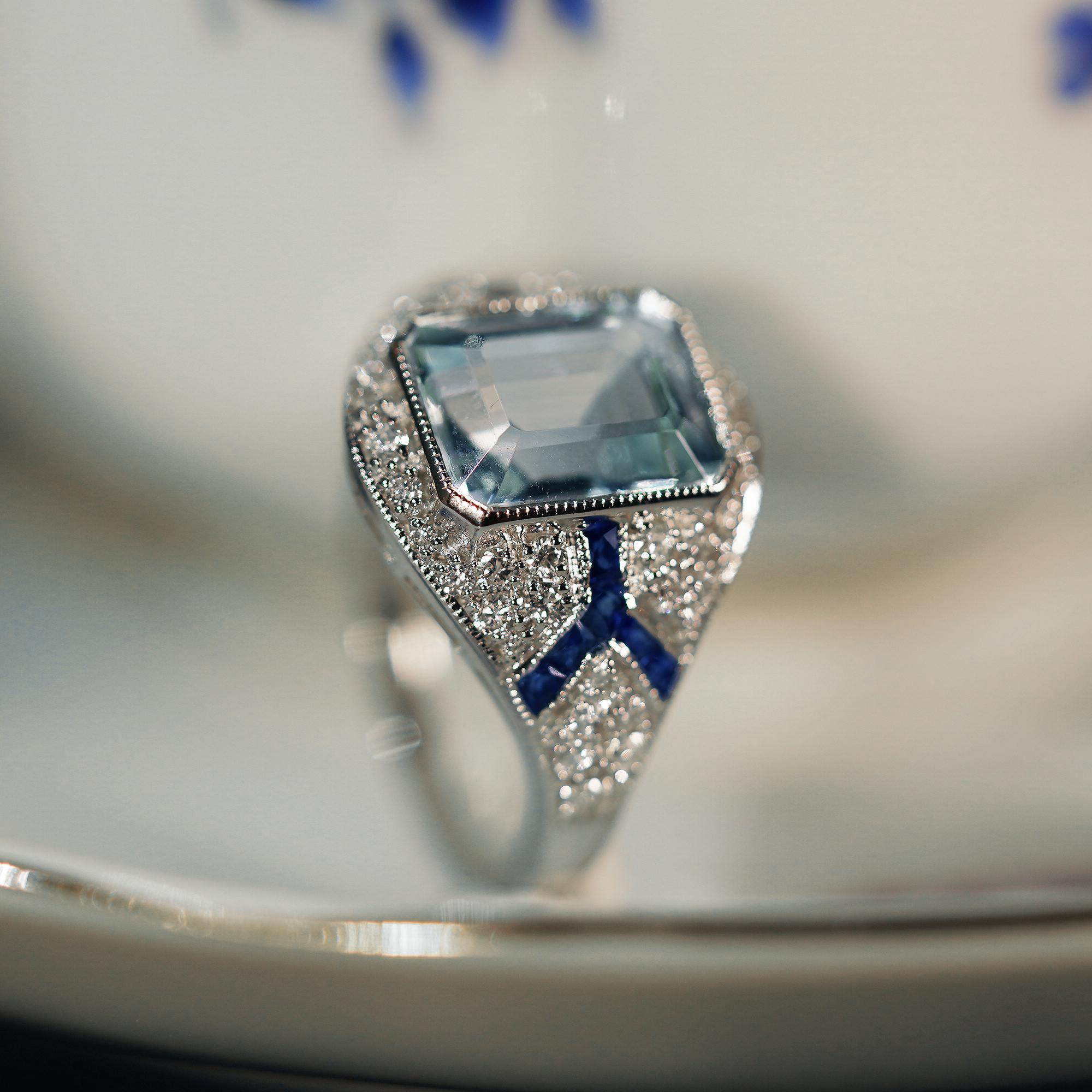 A gorgeous emerald cut aquamarine is presented in classic Art Deco style. The glorious light blue  gemstone radiates from within a sparkling halo of bright round brilliant cut diamonds punctuated east and west with two lines of French cut blue