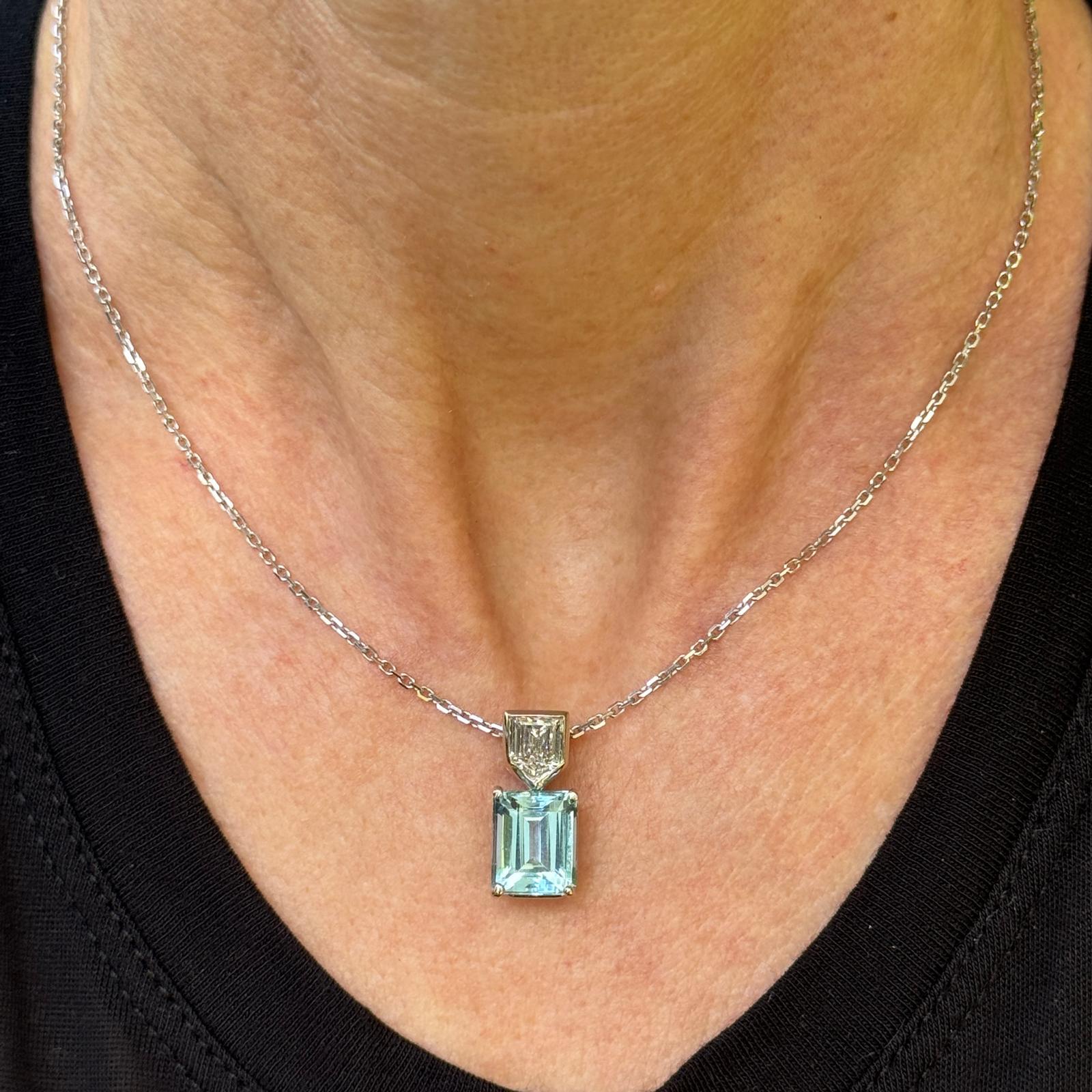 This elegant 14 karat white gold pendant showcases a stunning emerald-cut aquamarine of approximately 5.25 carats, radiating a captivating sea-blue hue. Stacked above the aquamarine is a unique shield-cut diamond, weighing approximately 0.80 carats,