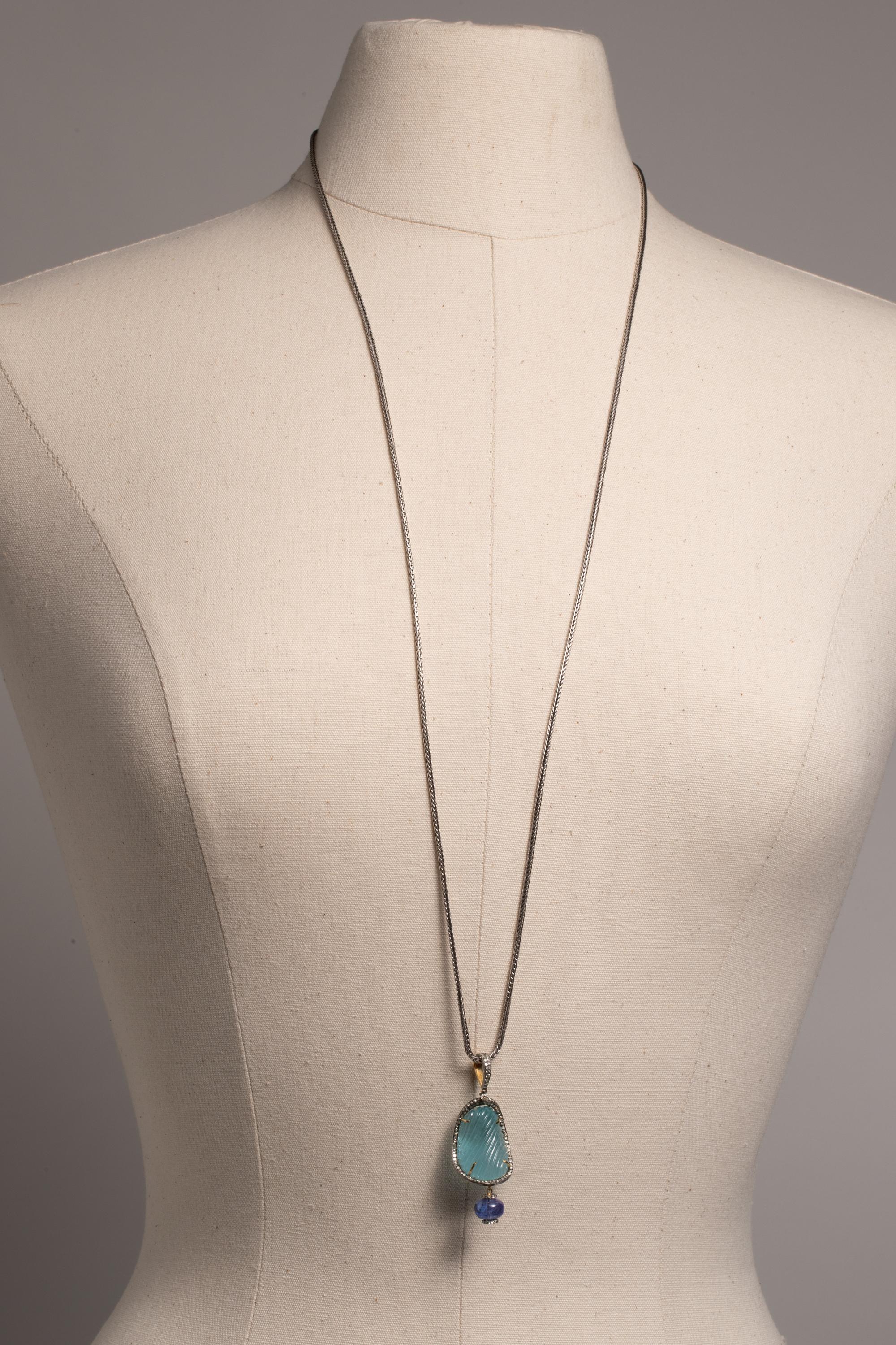 A long pendant necklace with a large hand-carved aquamarine bordered with round-cut, pave`-set diamonds and a diamond bail.  Round tanzanite bead bordered with diamond rondelles dangles underneath. Carat weight of the aquamarine is 37.88; the