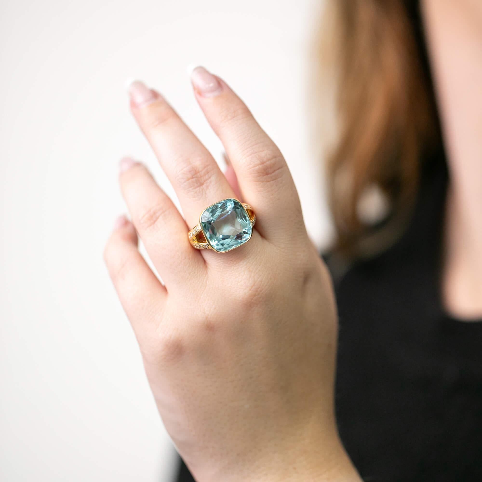 A late 20th Century hand-made ring by Anne Middleton is crafted in 18k yellow gold and features a bezel set cushion cut aquamarine with diamond set split shoulders upon a wide band. Stamped with the 750 hallmark and makers mark AM for Anne Middleton