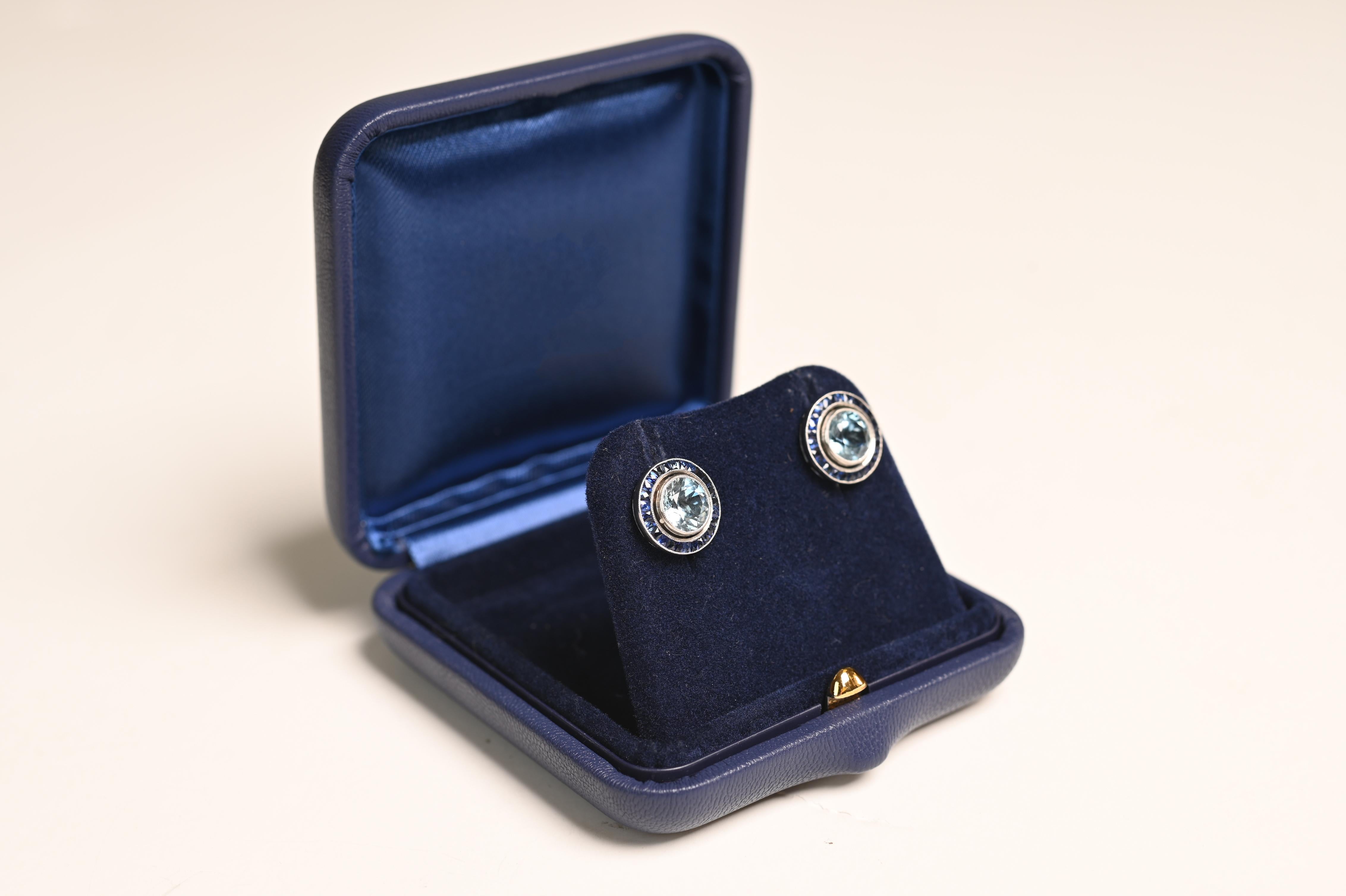 Cette paire de boucles d'oreilles exquise met en valeur deux aigues-marines rondes vibrantes, chacune possédant une saturation et une clarté remarquables. Les aigues-marines sont délicatement encadrées par un halo de saphirs bleus naturels taillés à