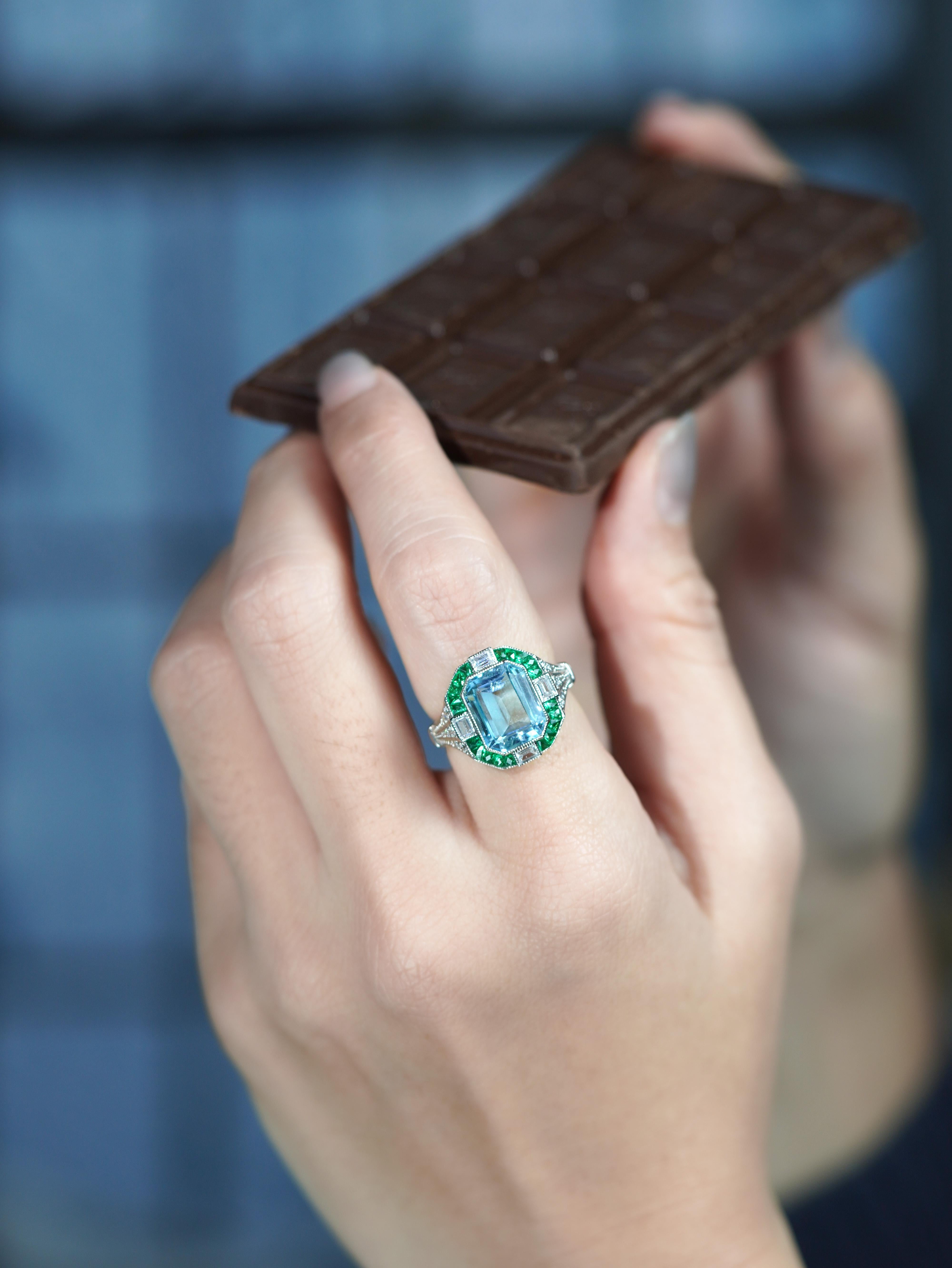 Gracieuse et captivante, cette bague de style Art déco met en valeur la beauté sereine d'une aigue-marine lumineuse en son centre. Encadré d'un halo d'émeraudes vibrantes de taille française et de diamants étincelants, le design rayonne d'une