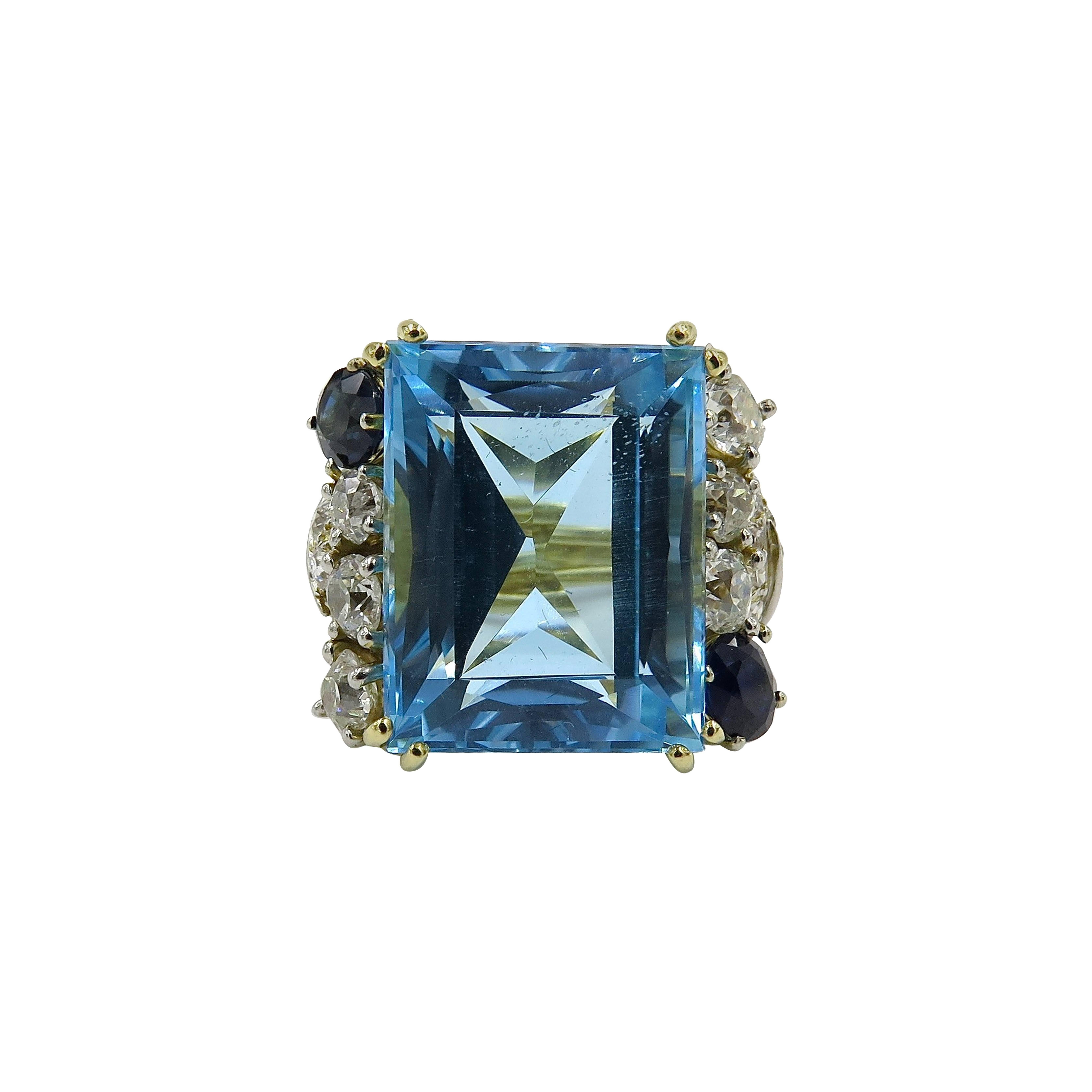 An 18 karat yellow gold, blue topaz, sapphire and diamond ring. Retailed by Milton Schepps. Set with a rectangular cut aquamarine, weighing 32.91 carats, enhanced by with old European cut diamond and sapphire accents pave shoulders, further enhanced