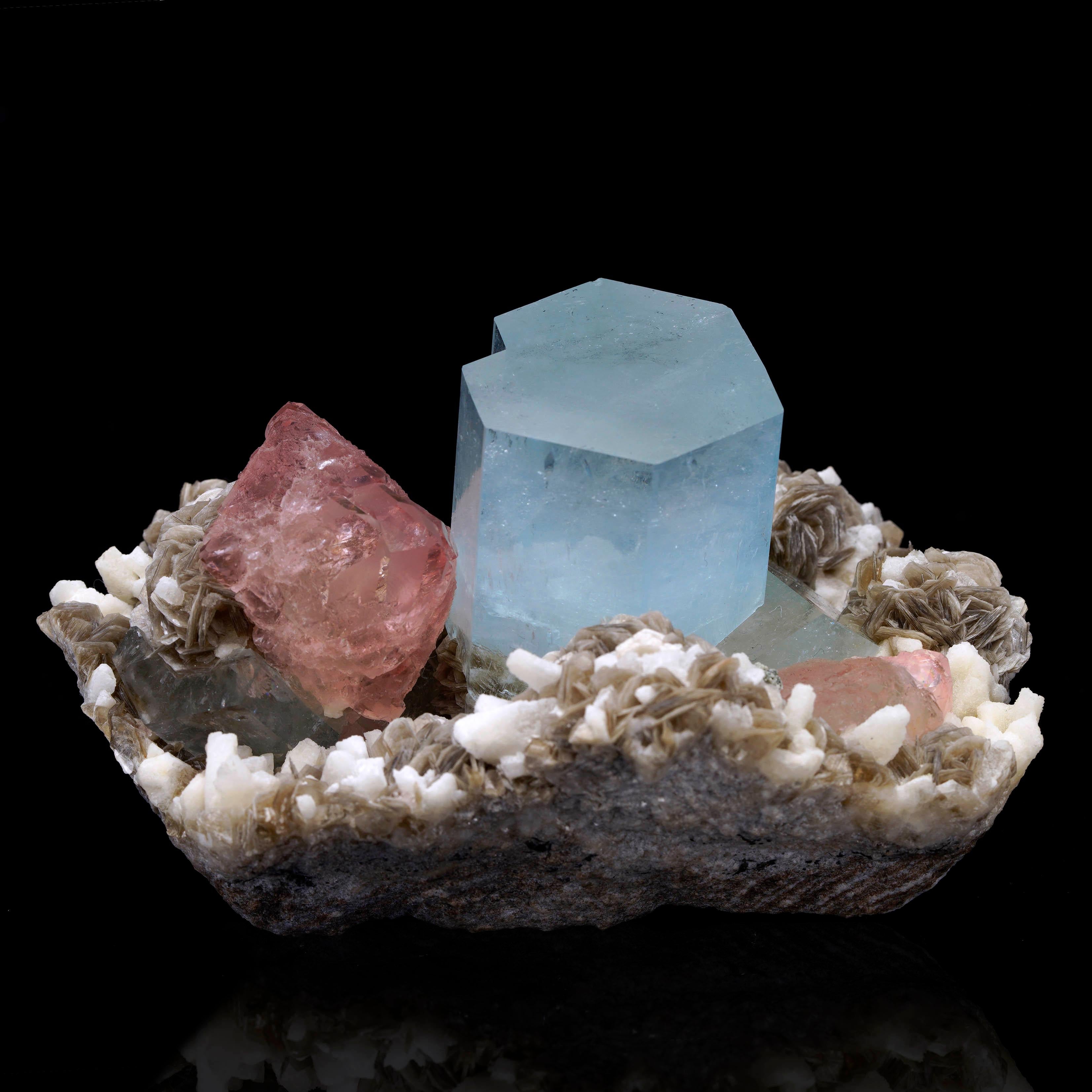 Un grande cristallo di acquamarina incontaminato sorge da un letto di muscovite argentata. I cristalli di fluorite rosa, tra cui un grande cristallo profondamente pigmentato a sinistra del centro che accentua il cristallo di acquamarina, contrastano