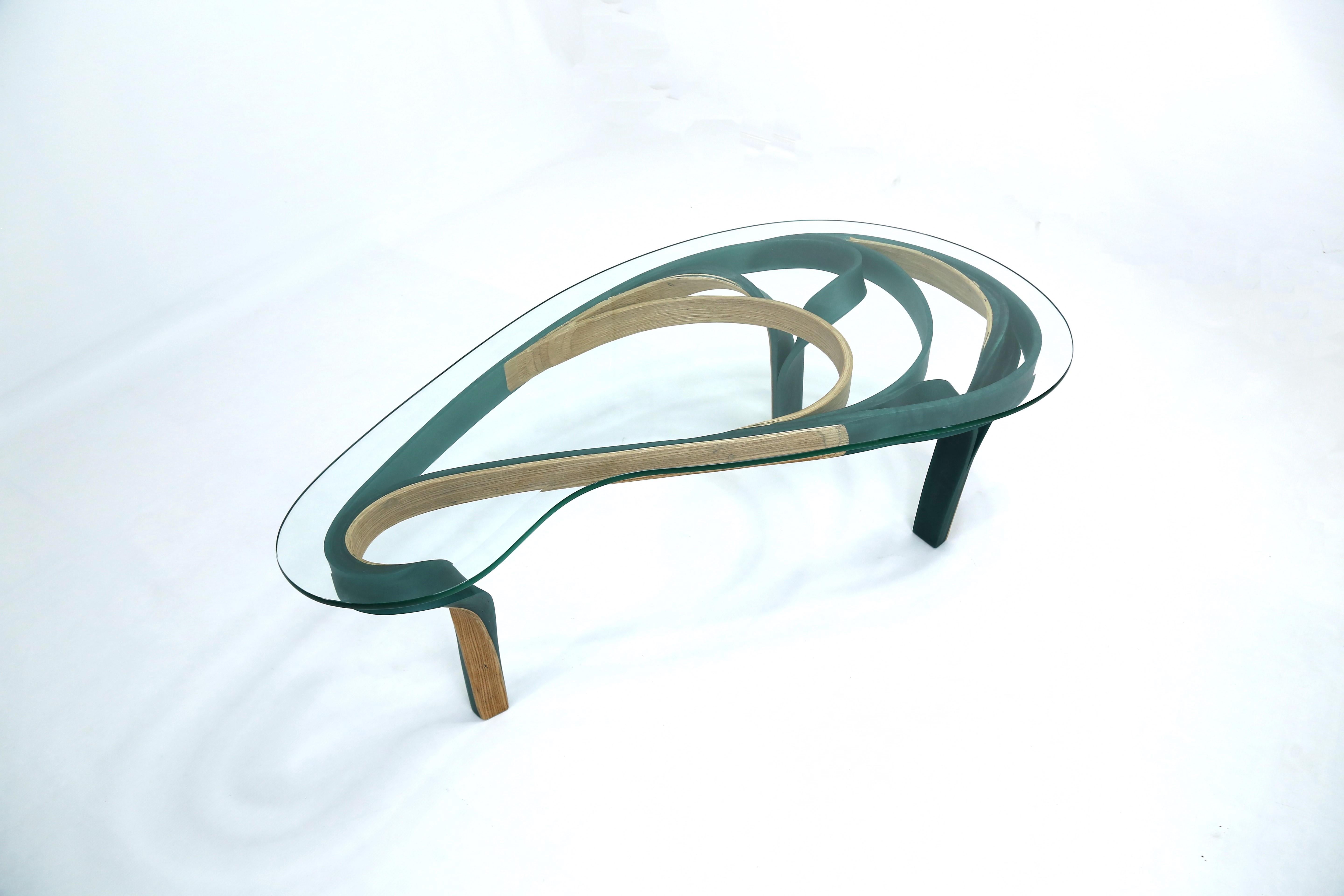 The Aquila Table has a bevelled glass top which is supported by a resin and ash wood frame in a paizlee shape which flows organically to create one of the supports. The other two supports stem smoothly from the frame and swirl towards the floor. The