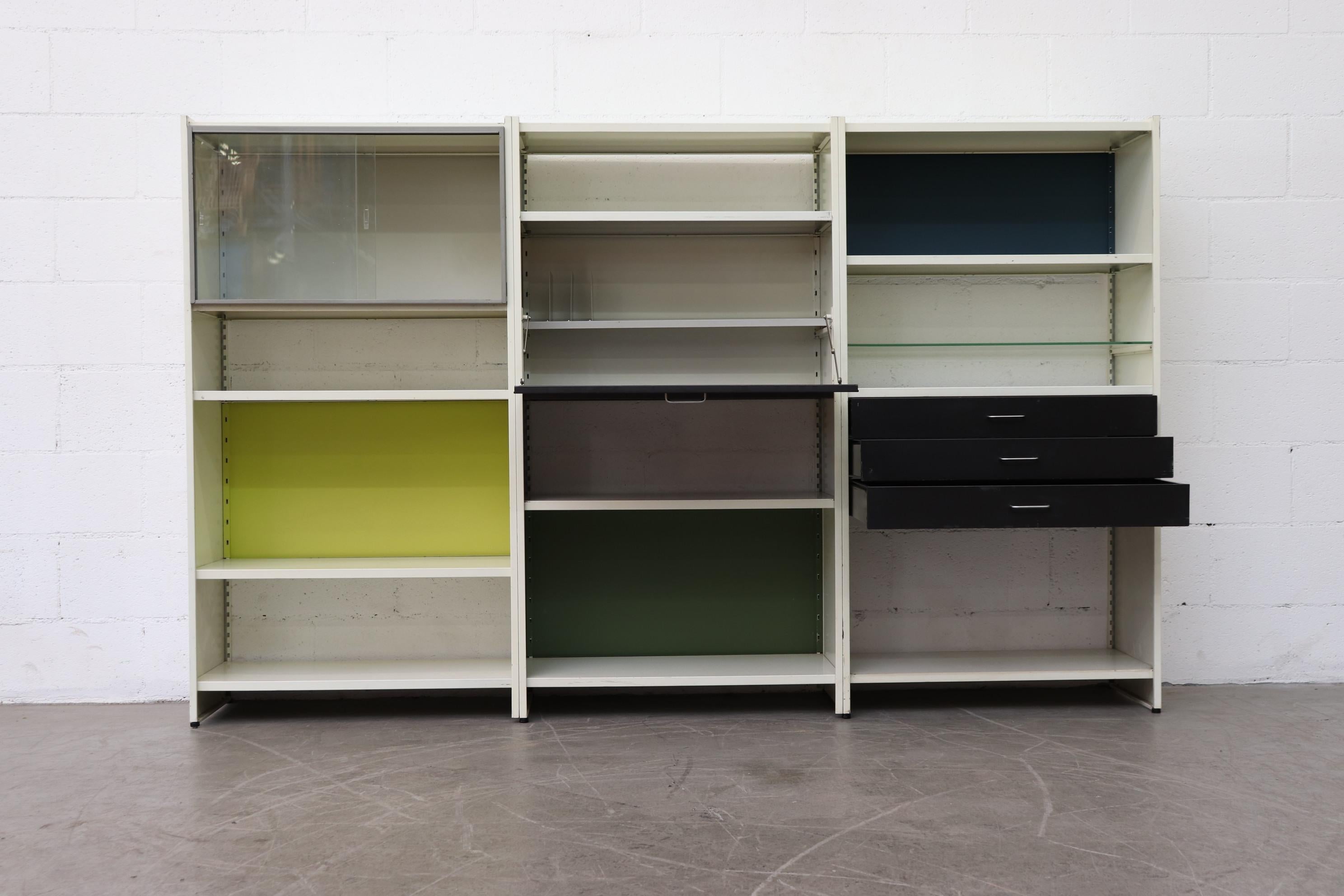 Large Gispen 3 section off-white metal industrial shelving and storage unit with drop down desk, 3 lower stacked drawers, cabinet with sliding glass doors and a glass shelf. Enameled metal panels in navy, bright yellow and green livens up the unit