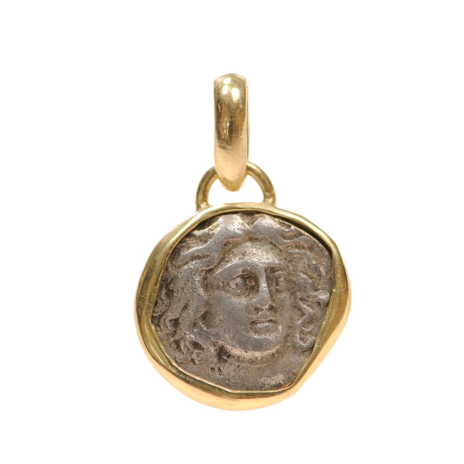 An AR Drachm Silver [coin] minted at Caria, Rhodes, Circa 205-190 BC, under the Magistrate Teisylos. There is simple and elegant 18 karat gold trim and bail around the coin. On the obverse side, is the 3/4 profile of Helios. On the reverse side,
