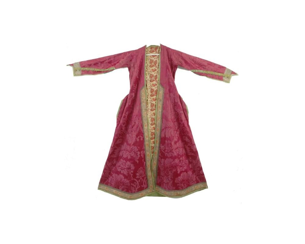 An antique Persian Iranian Safavid silk robe. The robe features a bold crimson base adorned with intricate floral patterns, complemented by ornate silver and golden embellishments. The robe exhibits detailed floral motifs woven into the fabric, with