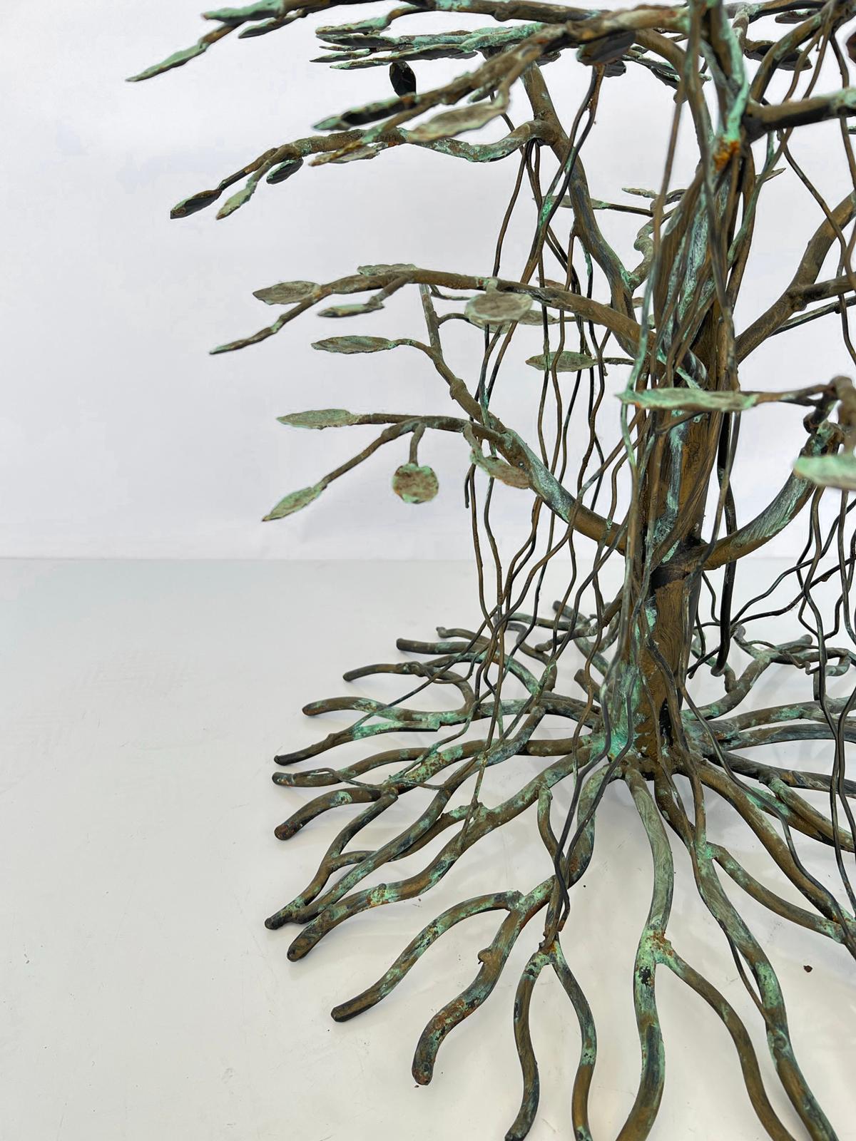Signature arboreal accent table, having a round top of glass, on a base of welded bronze with a verdigris finish, fashioned as a stylized banyan tree. Its central trunk affixed with leafy branches, with ariel and ground roots, The table can work as