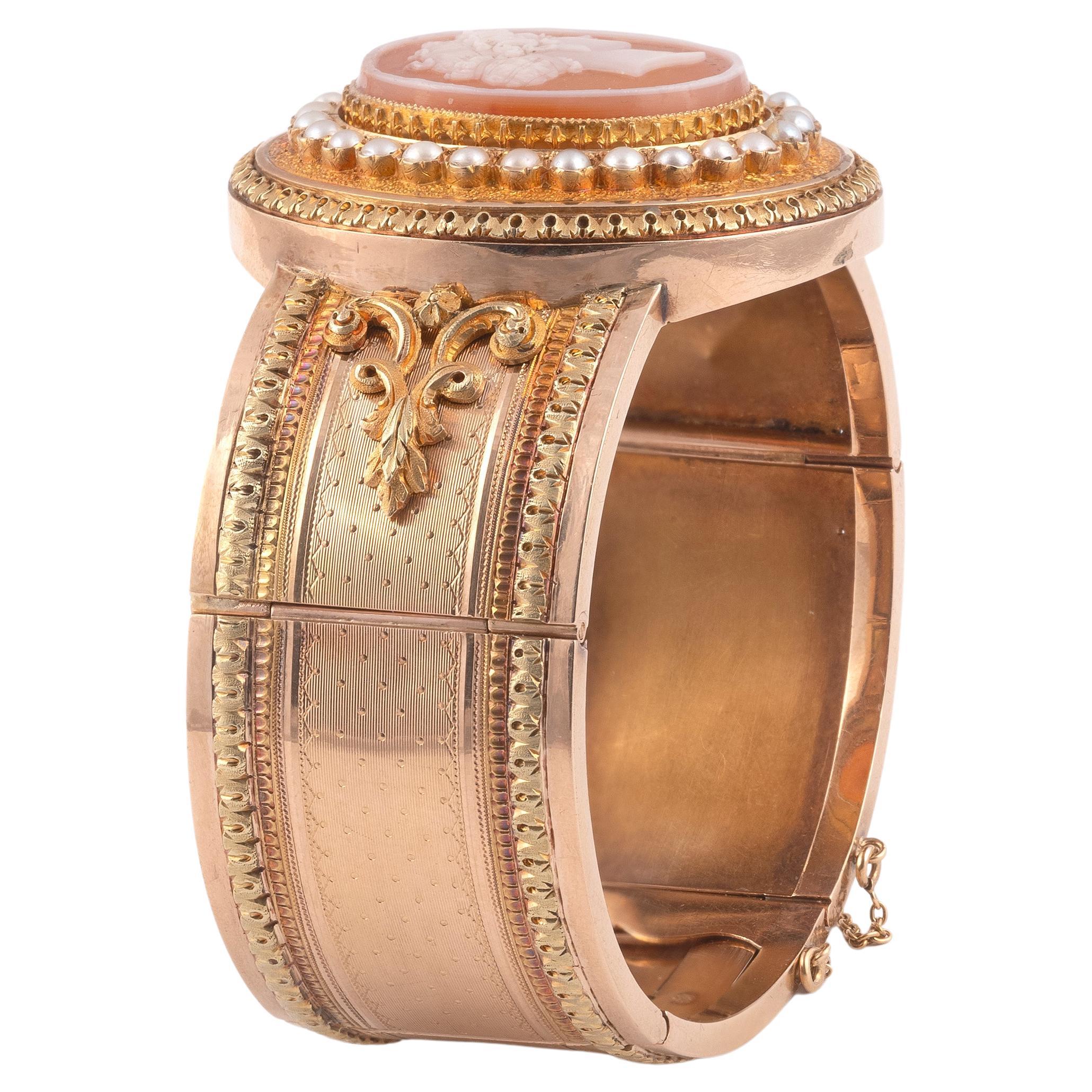 Ha la forma di un bracciale rigido apribile decorato al centro con un cammeo ovale su corniola a due strati che rappresenta una donna elegante con orecchini in una cornice di mezze perle pregiate. Cornice in oro 18 carati a tre colori, cesellata,